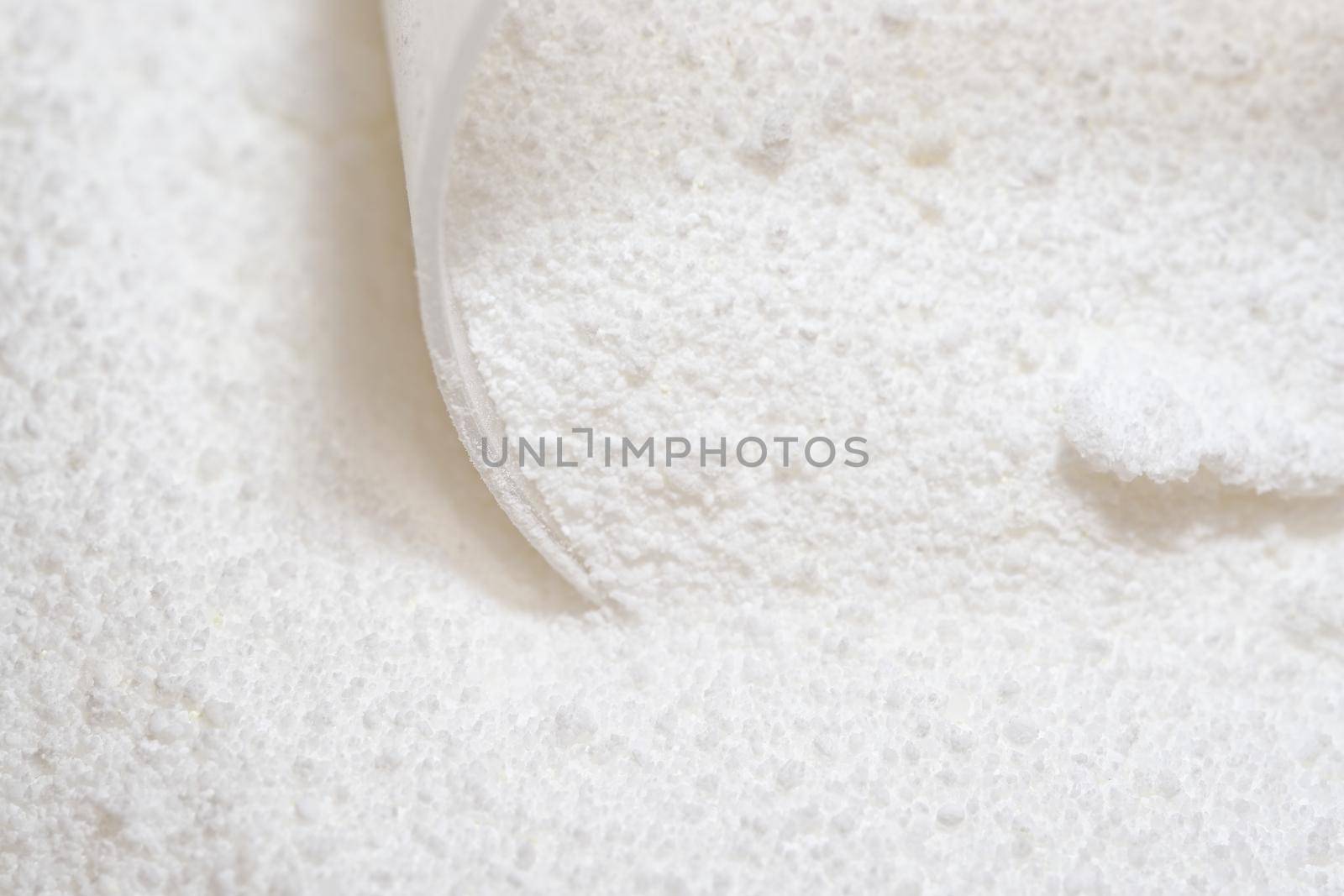 Pure white laundry detergent powder, clumped together and being picked up by a clear plastic measuring scoop. Bright image and super macro perspective
