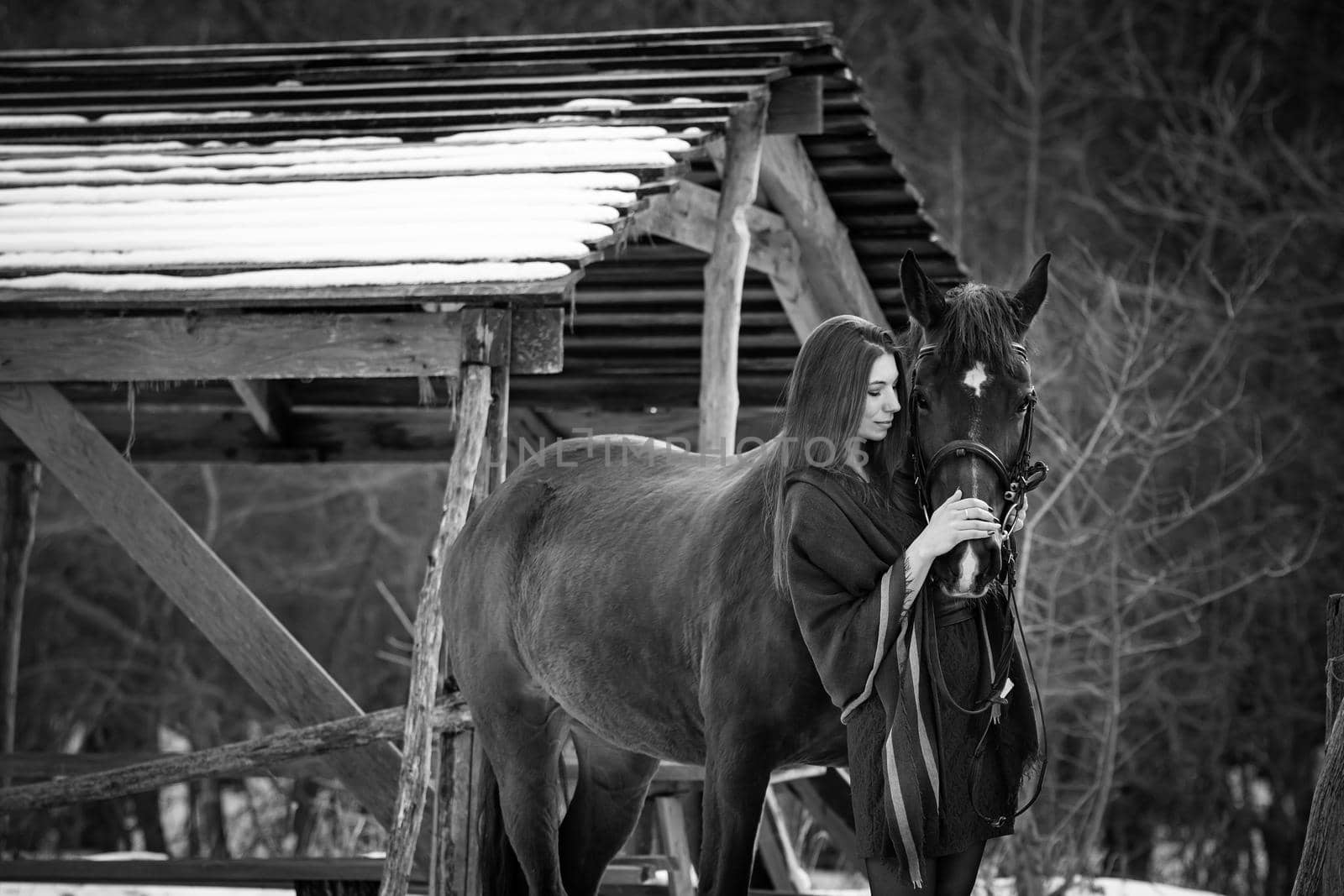 Beautiful girl hugs a horse near wooden buildings on a winter day, black and white photography