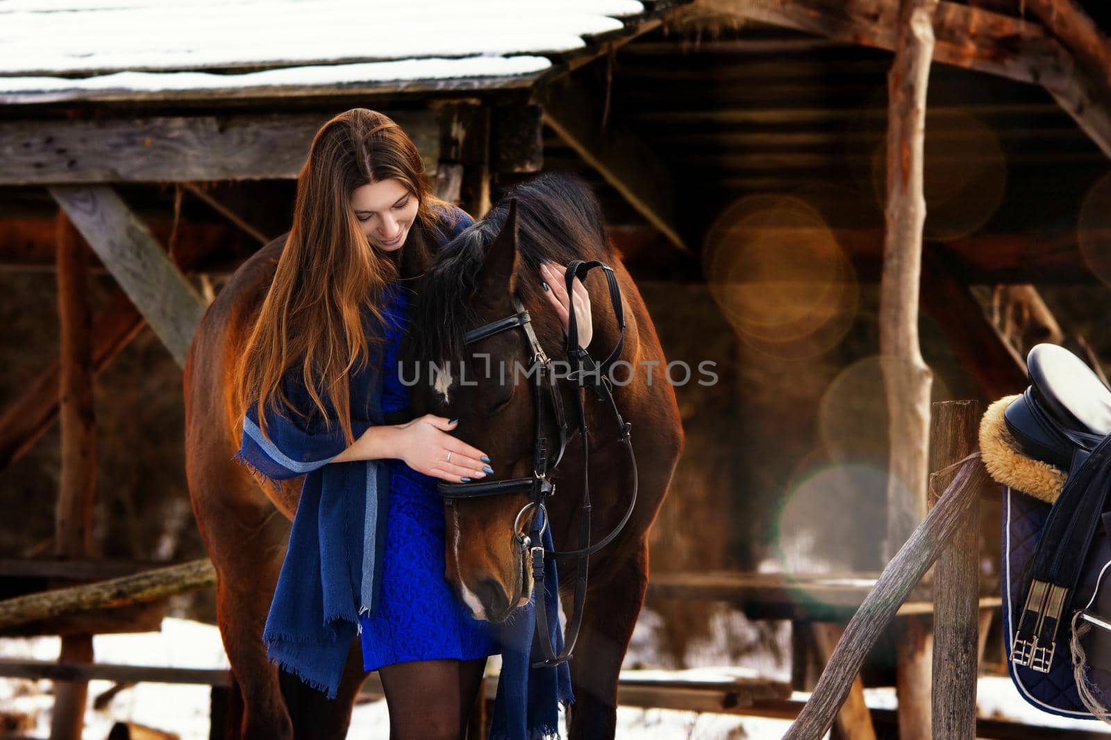 Beautiful girl in a blue stole hugs a horse near wooden buildings on a winter day by Madhourse