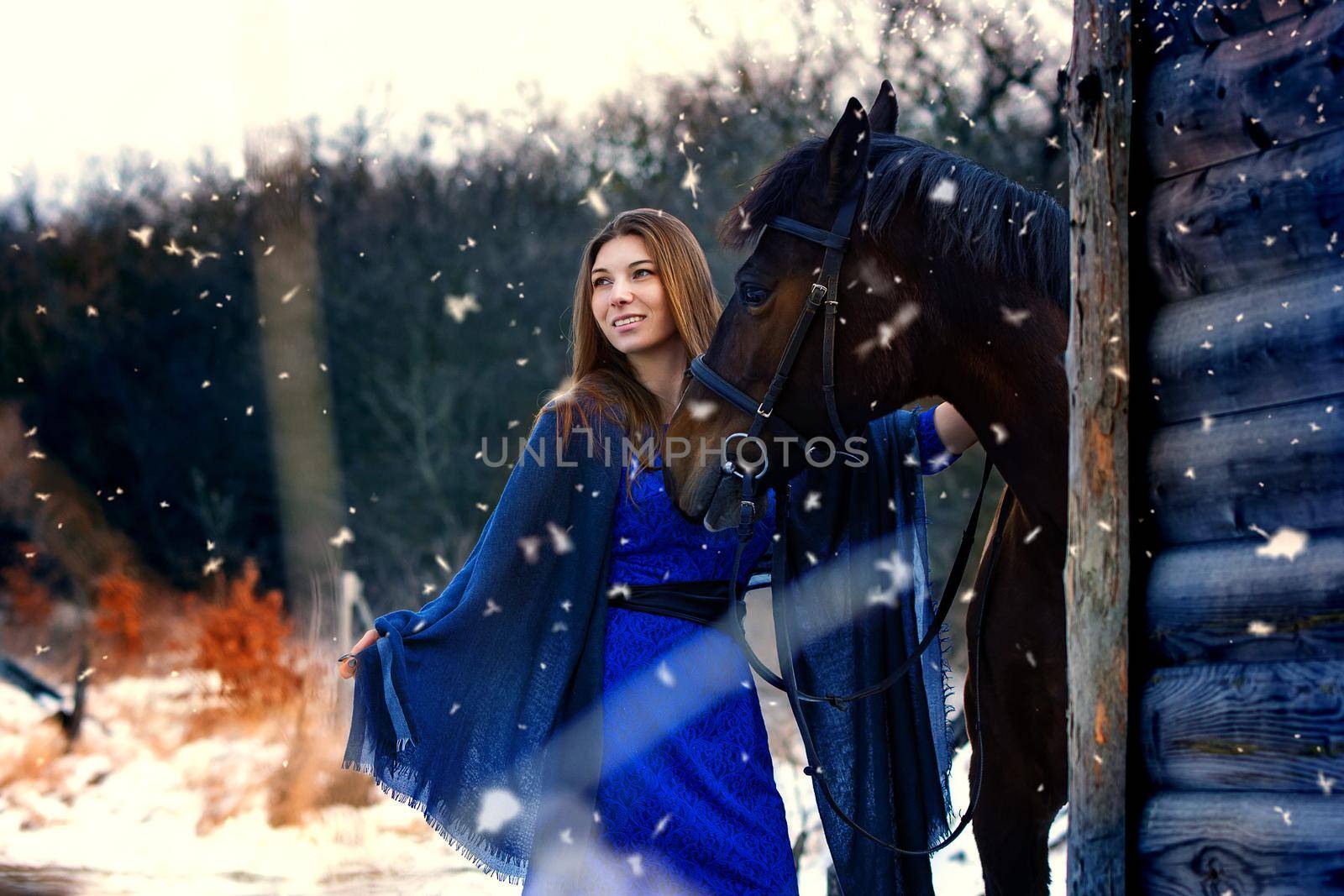 Beautiful girl in a blue stole stands next to a horse near wooden buildings on a snowy winter day by Madhourse