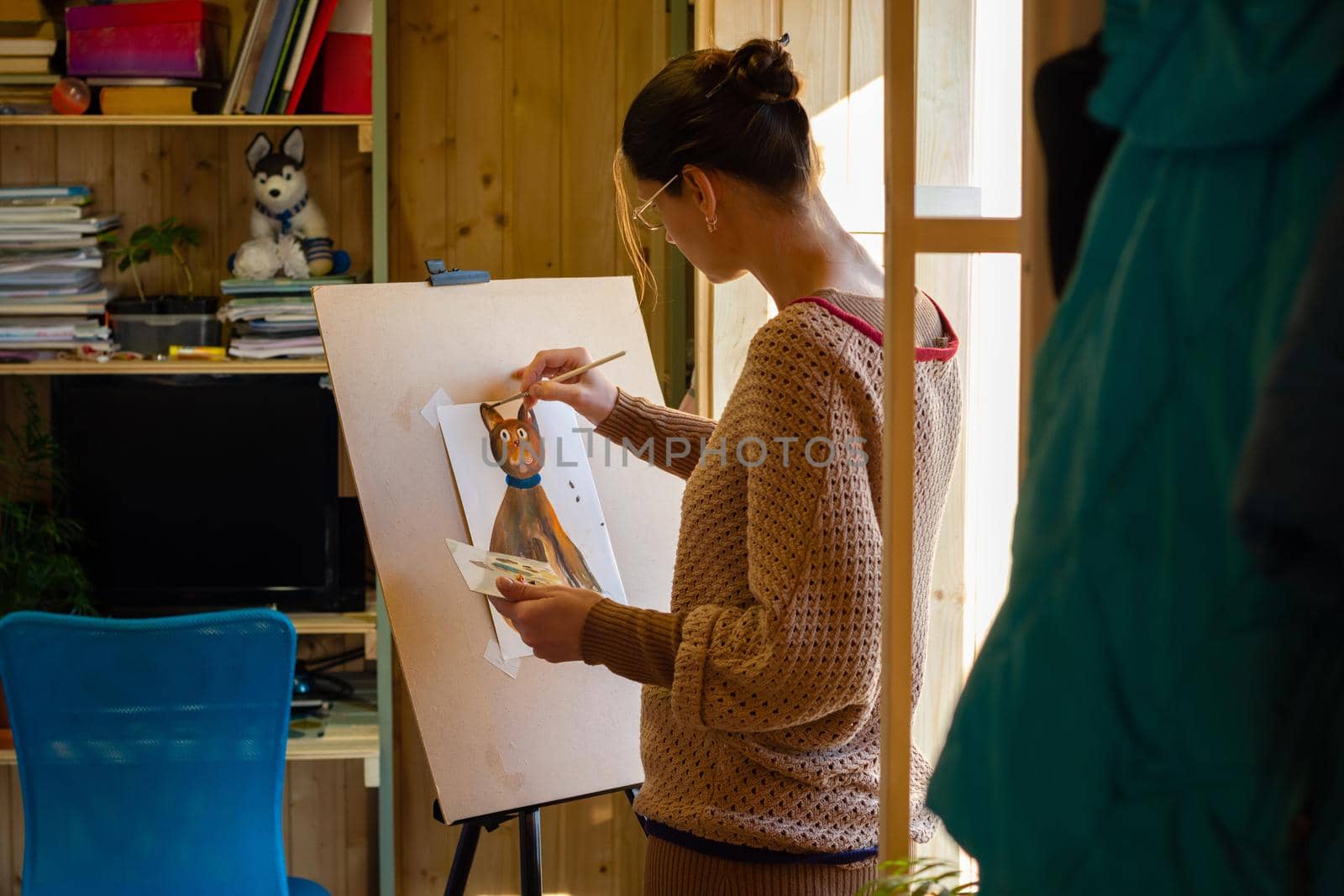 The artist paints a drawing of a cat with acrylic paints on an easel, in a small workshop by the window