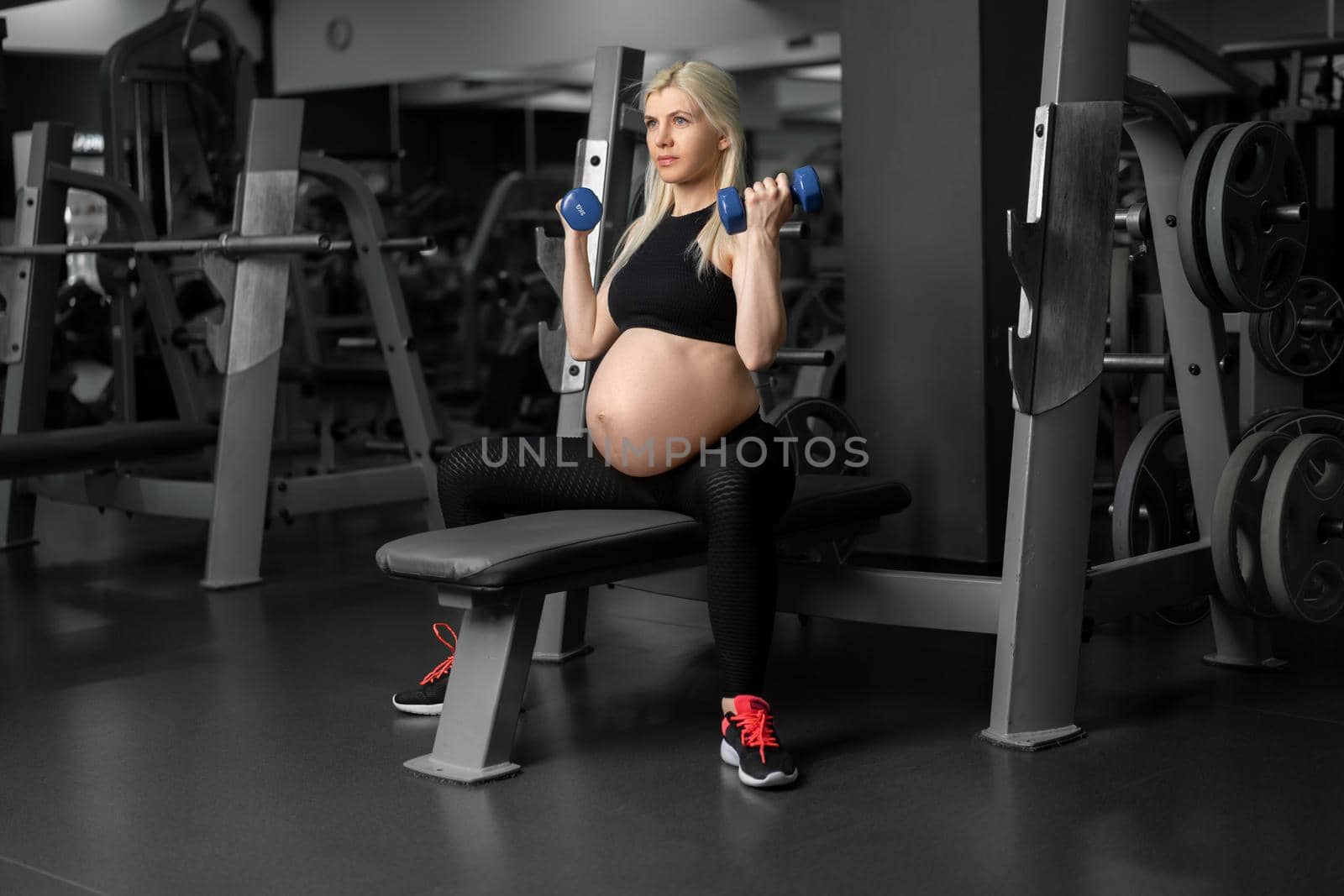 Pregnant woman lifting up dumbbells training biceps muscle at the gym sitting bench Pregnancy, healthy lifestyle, sport and fitness concept Caucasian female athlete workout training