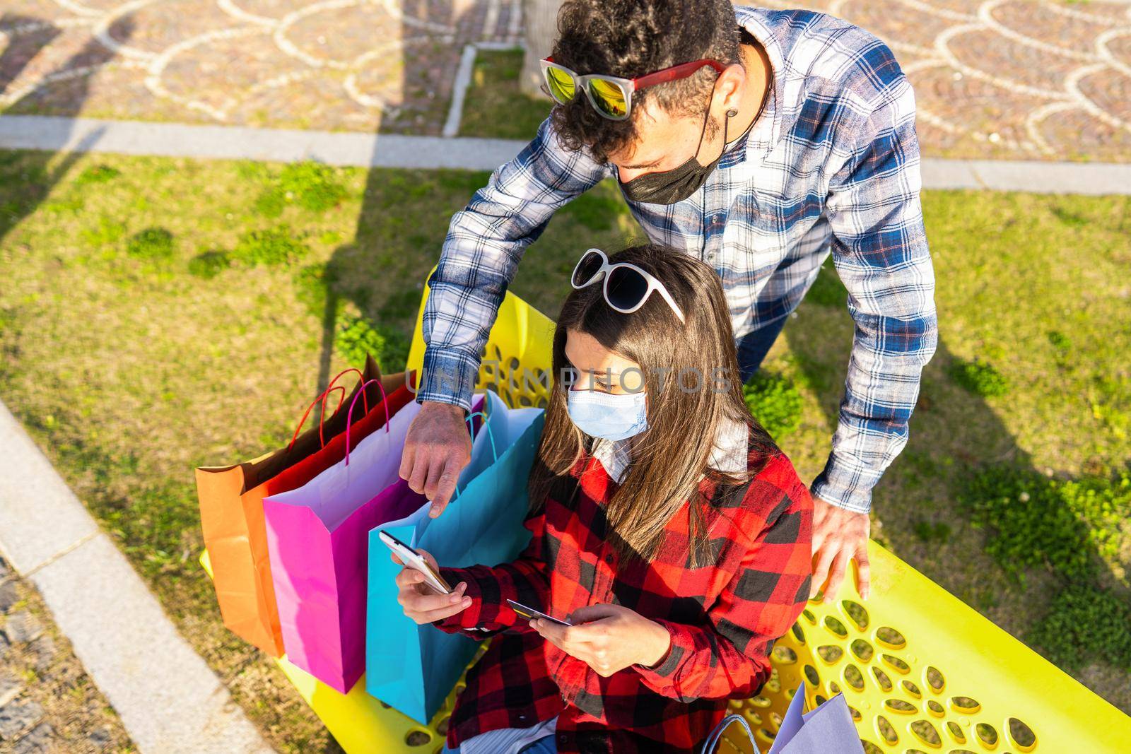 Young couple in protective mask using smartphone to check online shopping deal using internet mobile connection sitting on a city park bench among a lot of colored shopping bags. Girl with credit card by robbyfontanesi