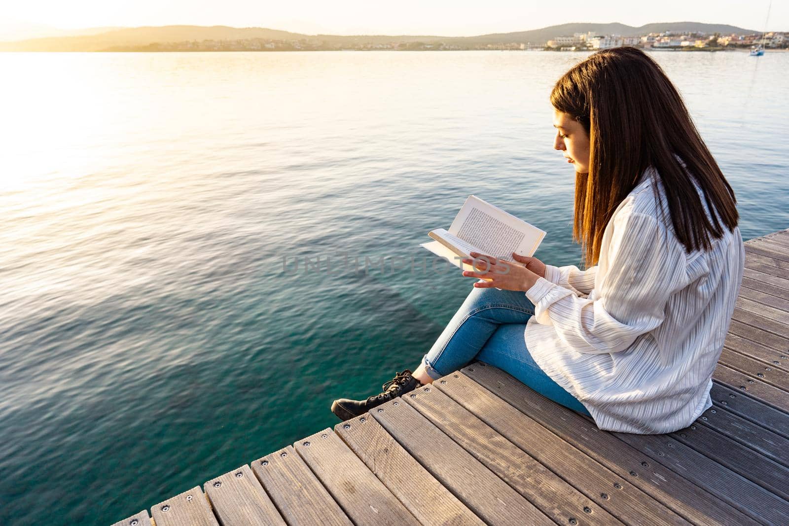 Young woman sitting on a pier at sunset or sunrise on transparent sea ocean water reading a book. Student girl learn university school lesson in nature. Bookaholic person having fun with romance tale