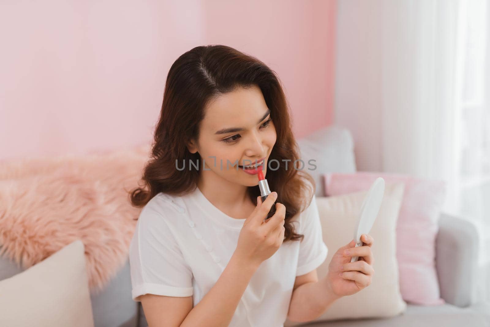 Smiling young beautiful asian woman fresh healthy skin looking on mirror and enjoying applying lips with red lipstick at home.facial beauty and cosmetic concept by makidotvn