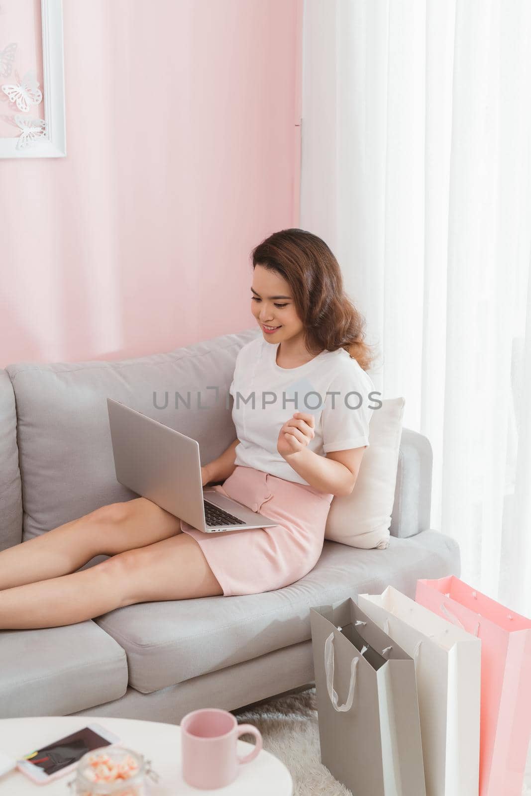 A happy woman shopping online with credit card from her modern home while sitting on sofa with laptop.