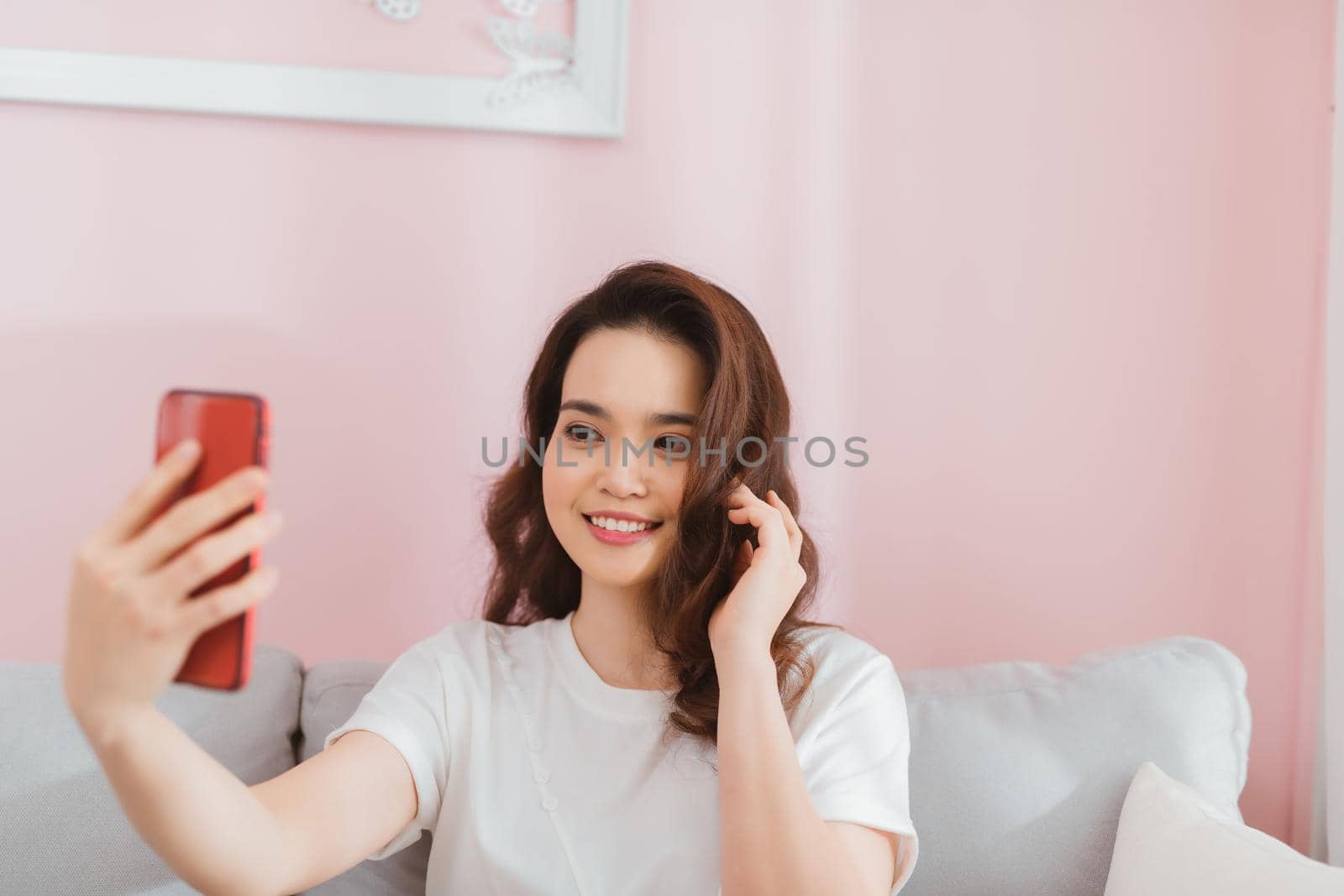 Attractive smiling young asian woman wearing casual clothes sitting on a couch, taking a selfie
