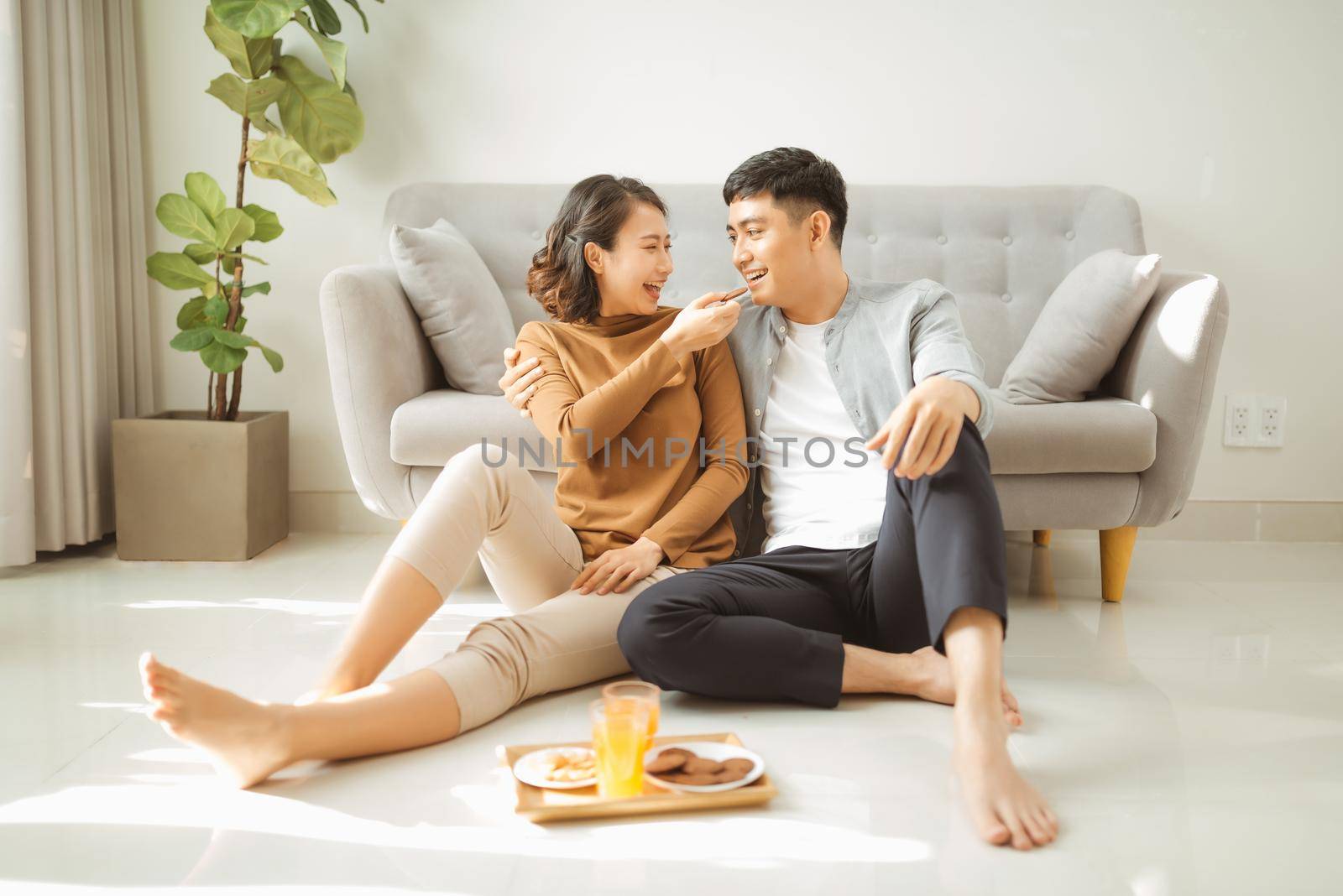 Relaxing at home. Happy young man and woman drinking hot beverage with sweet pastry. They are sitting at table on floor near couch and smiling