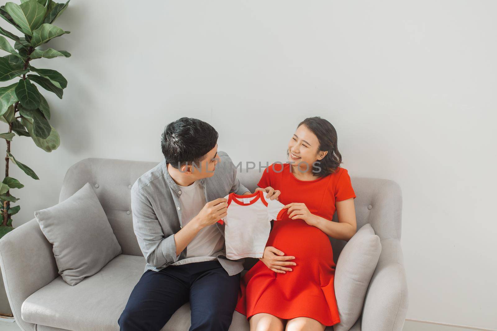 Young husband and a pregnant wife show their baby t-shirt when sitting on sofa.
