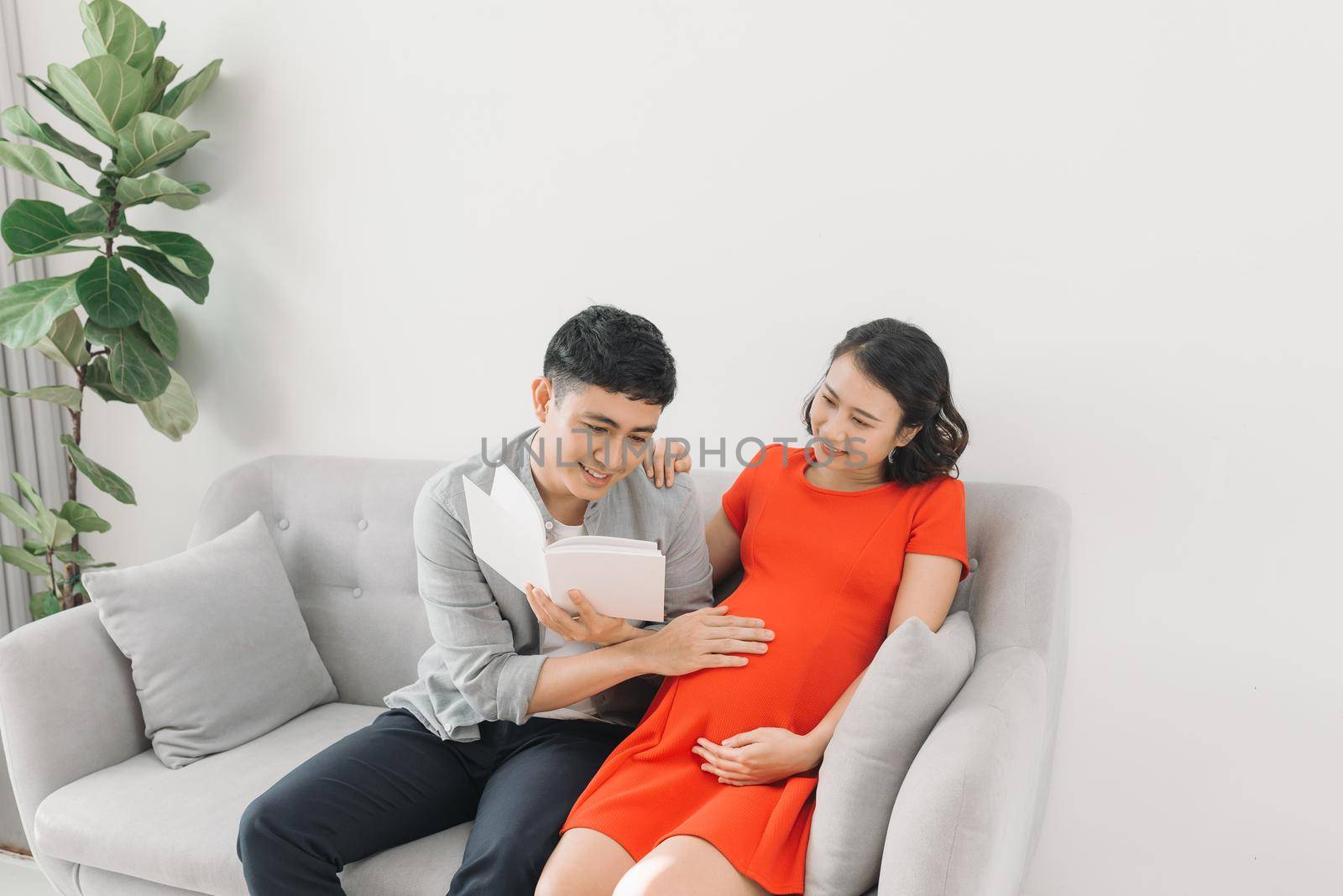 Asian pregnant woman and husband sitting on sofa and reading book