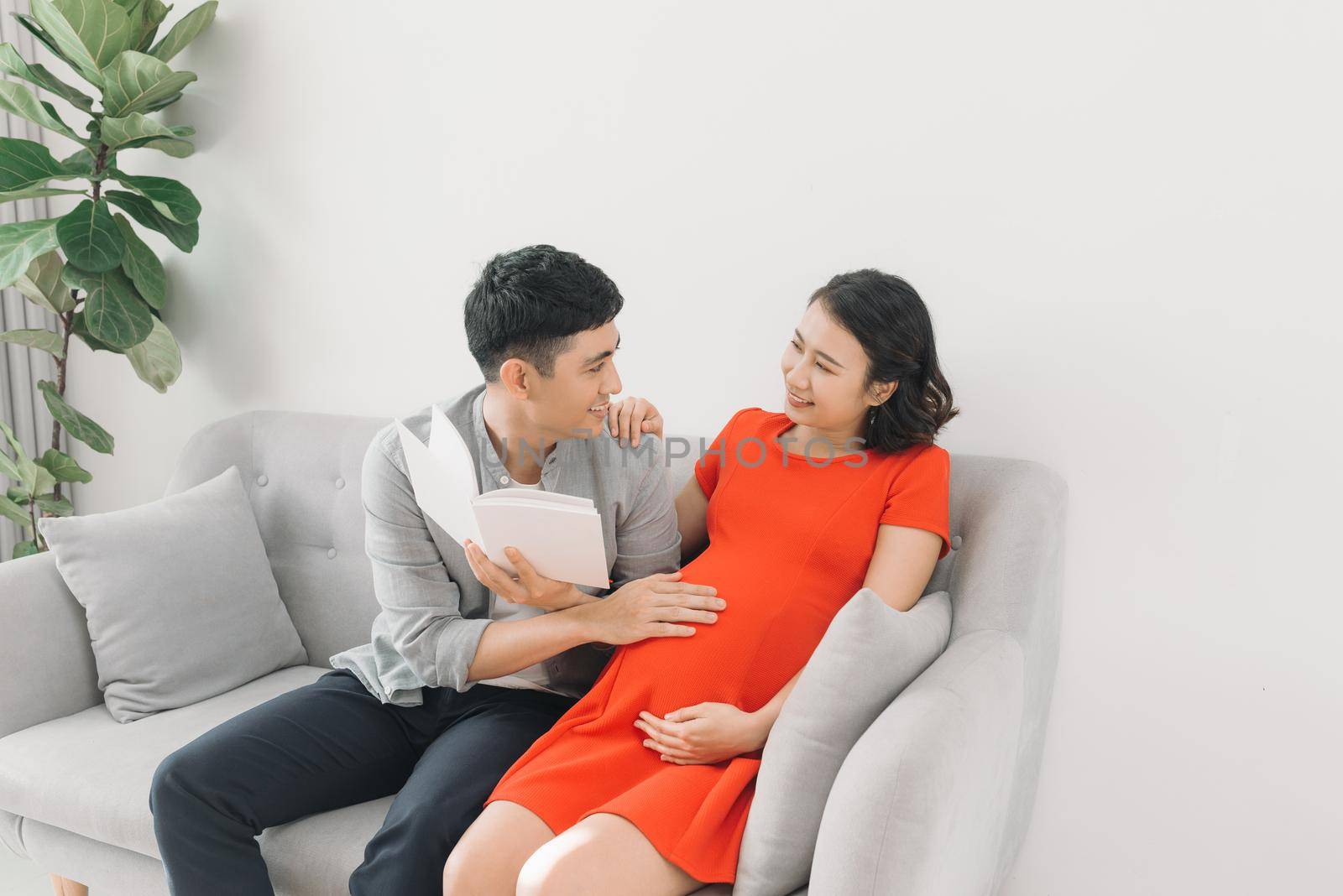 Asian pregnant woman and husband sitting on sofa and reading book