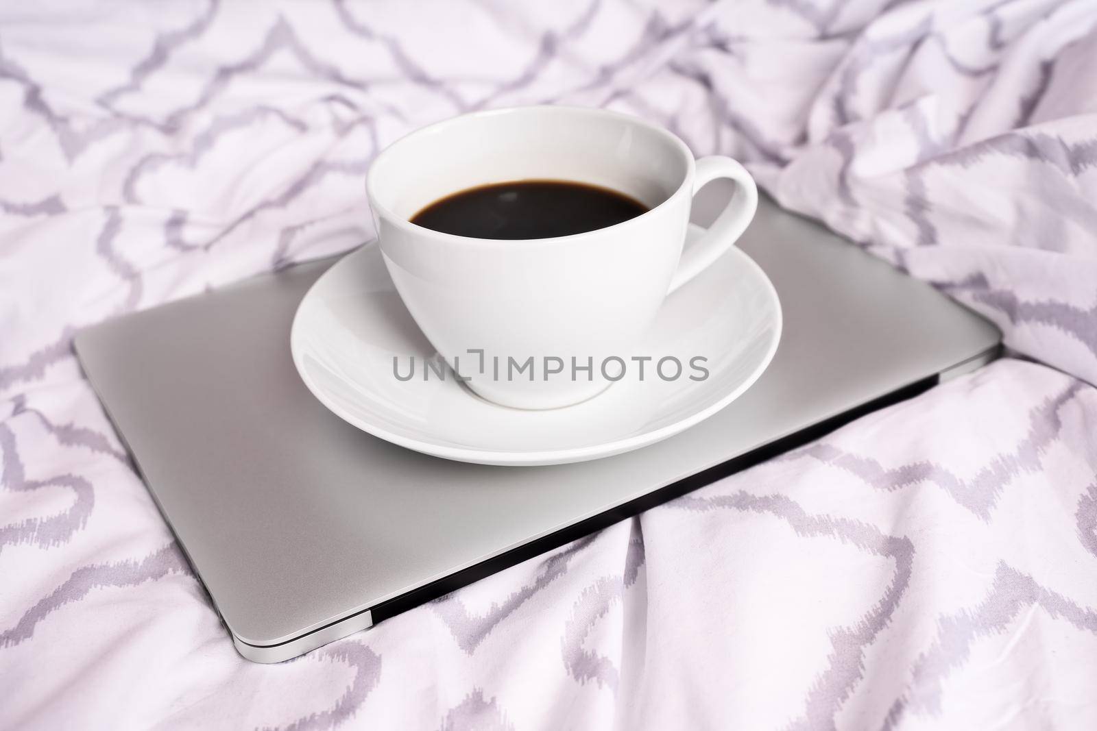 White cup of coffee on top of a laptop on bed with modern bed sheets. Work from home concept. Morning light. Mobile working concept.