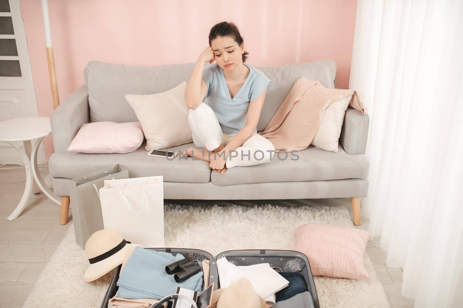 Young woman having trouble packing suitcase bag, sitting on sofa, hand on face concerned, not ready