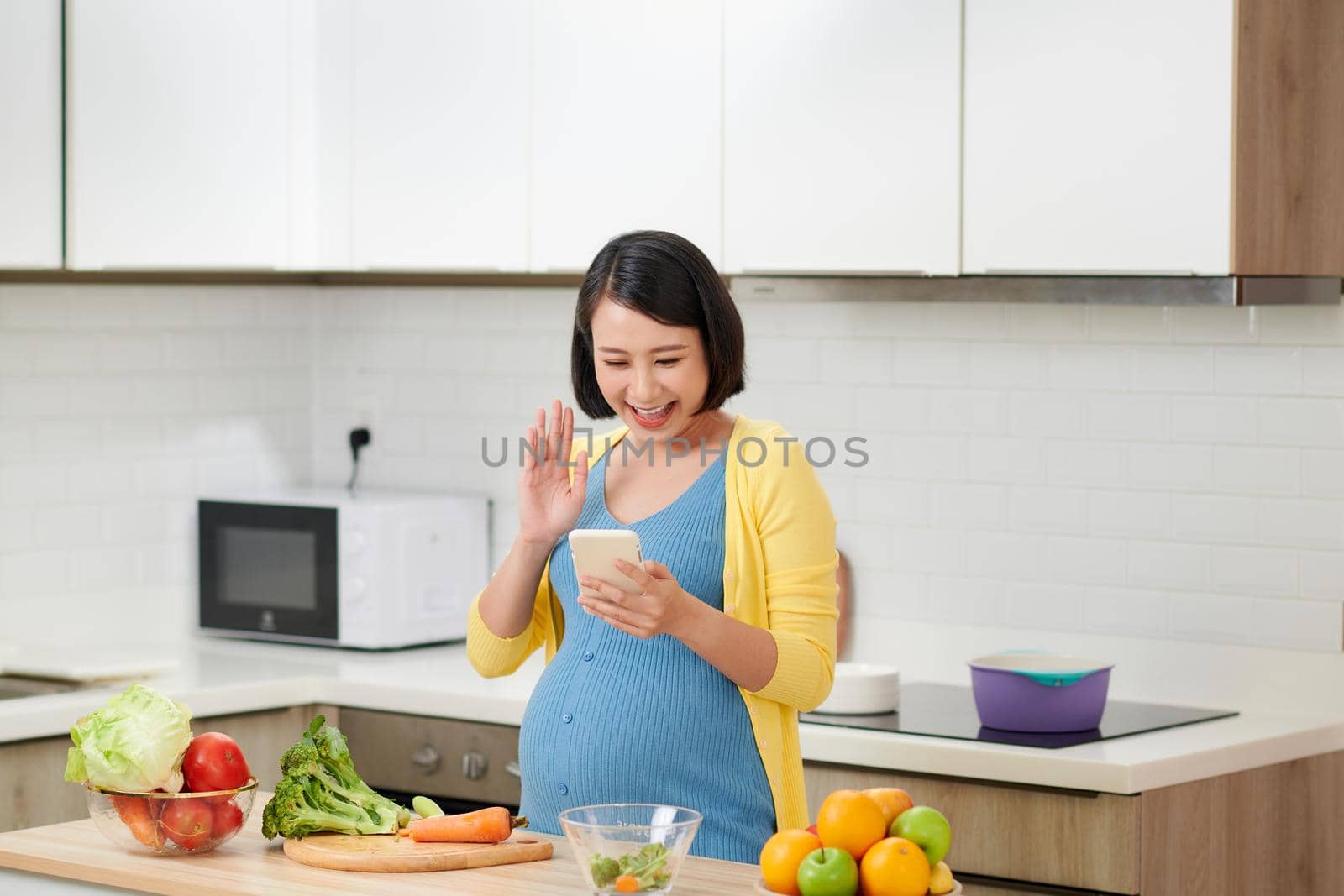 Happy pregnant woman calling on smartphone and having breakfast at home kitchen