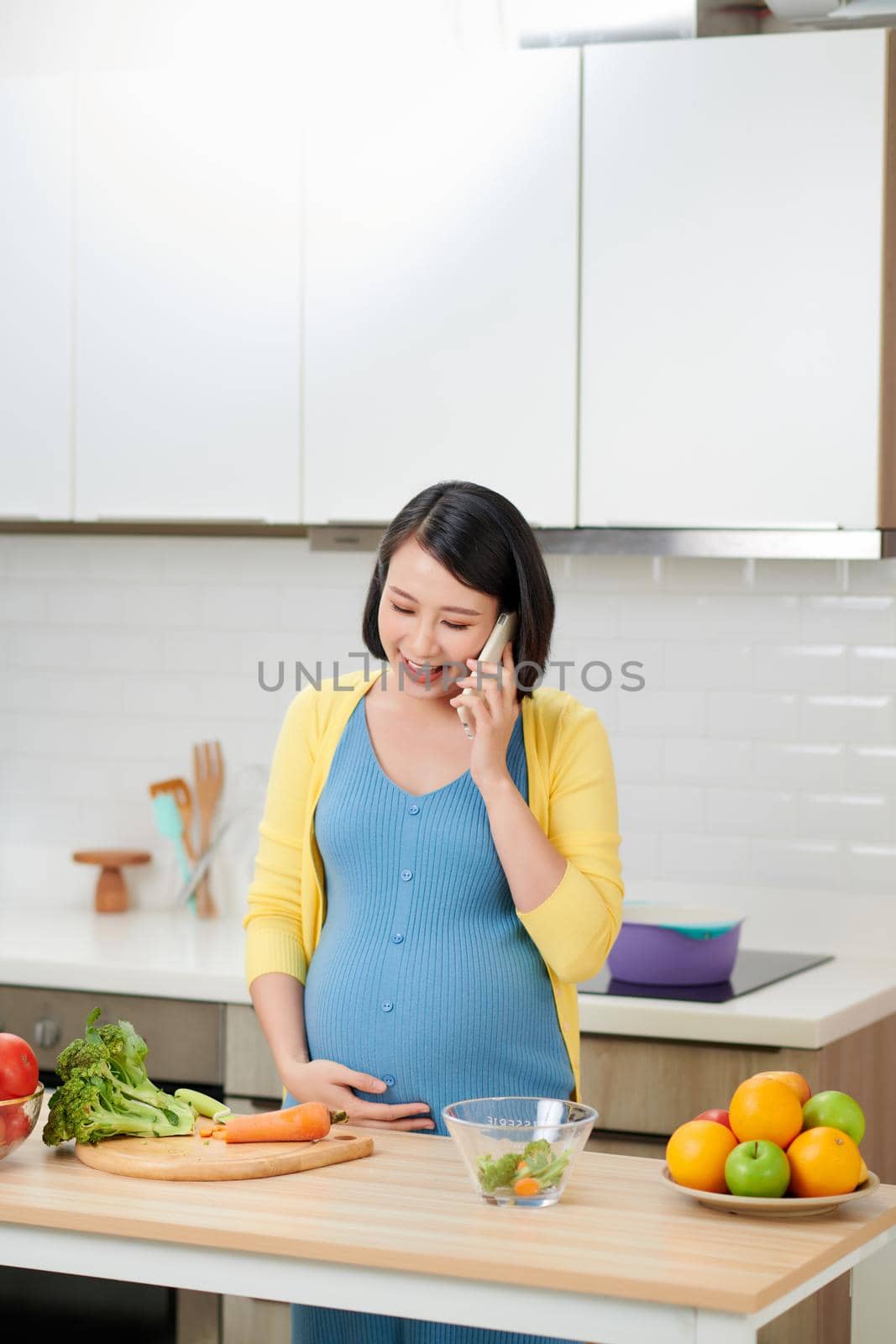 Happy pregnant woman calling on smartphone and having breakfast at home kitchen