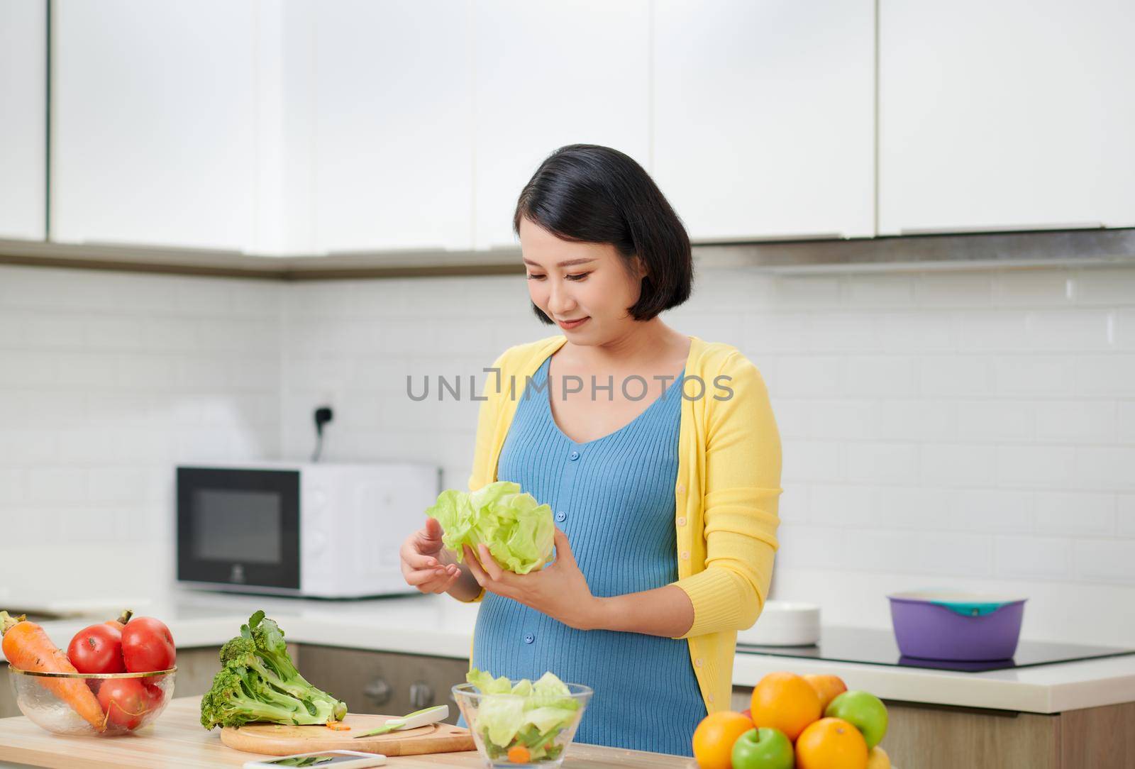 Happy pregnant woman cooking at home, doing fresh green salad, eating many different vegetables during pregnancy, healthy pregnancy concept