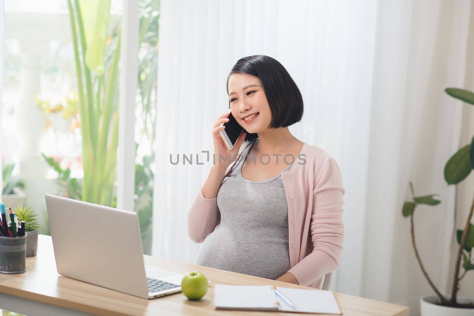 Pregnant businesswoman working with laptop and talking on phone in cozy bright office.