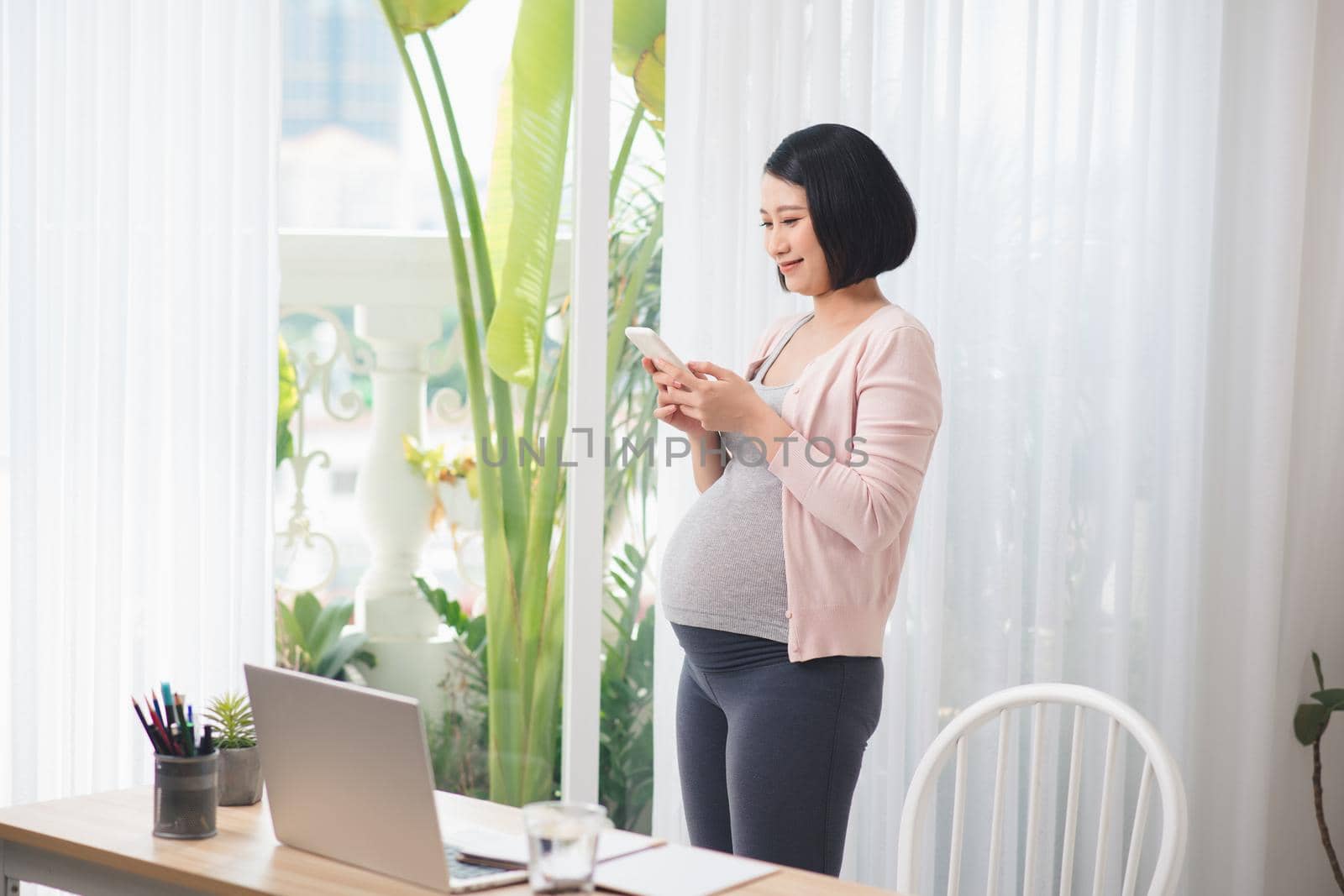 Young pregnant woman standing at window and reading text message on smartphone