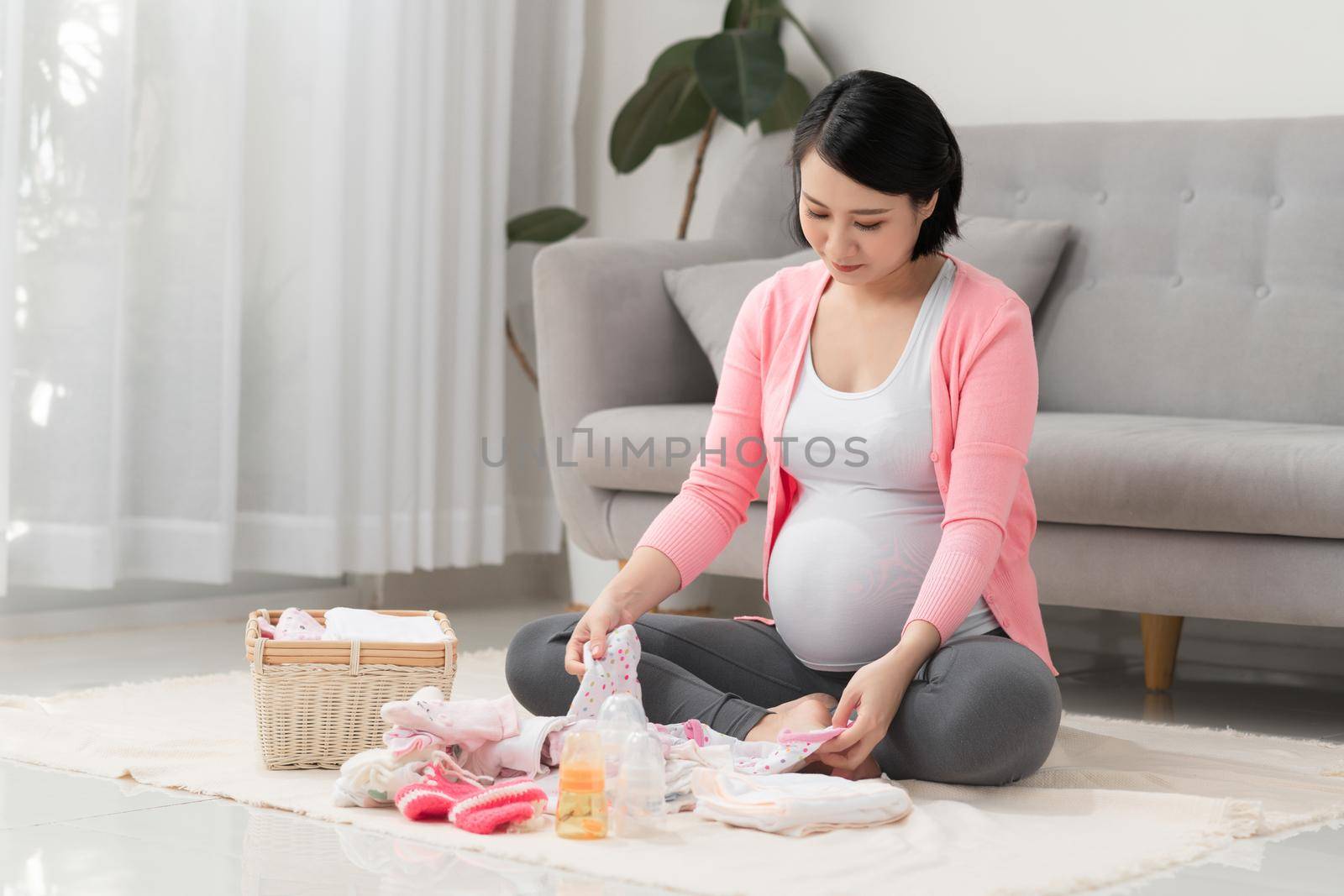 Young pregnant woman holds baby clothes on white background sitting at home on the floor
