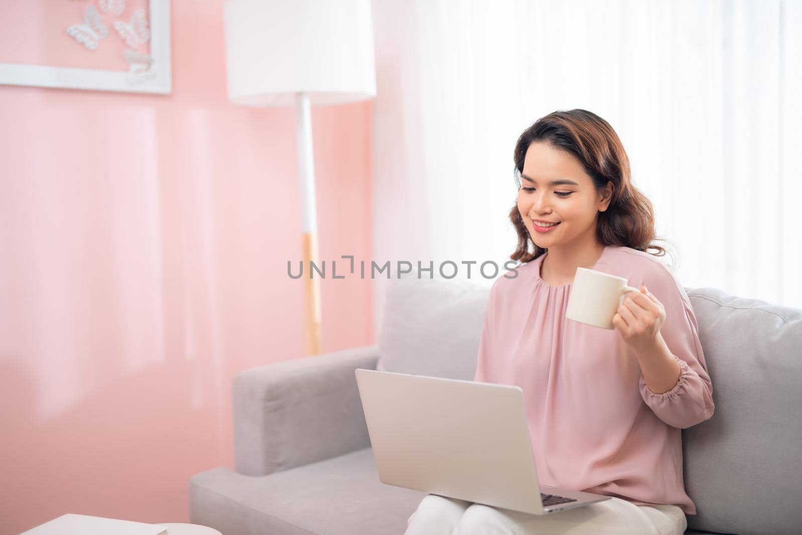 Attractive Asian female lifestyle using the laptop sitting on a couch and drinking coffee