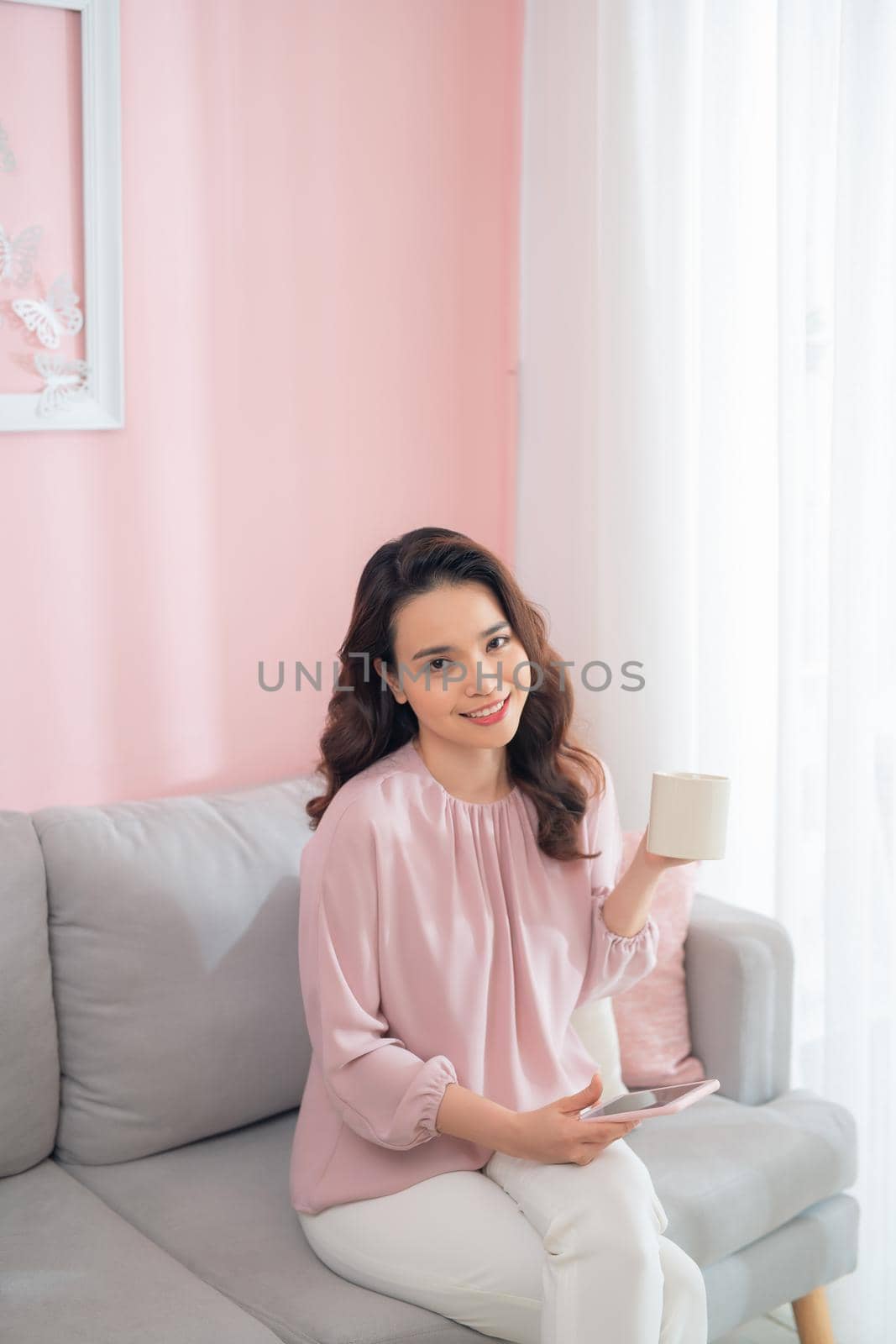 Attractive young Asian woman using phone and drinking coffee when sitting on sofa at home.