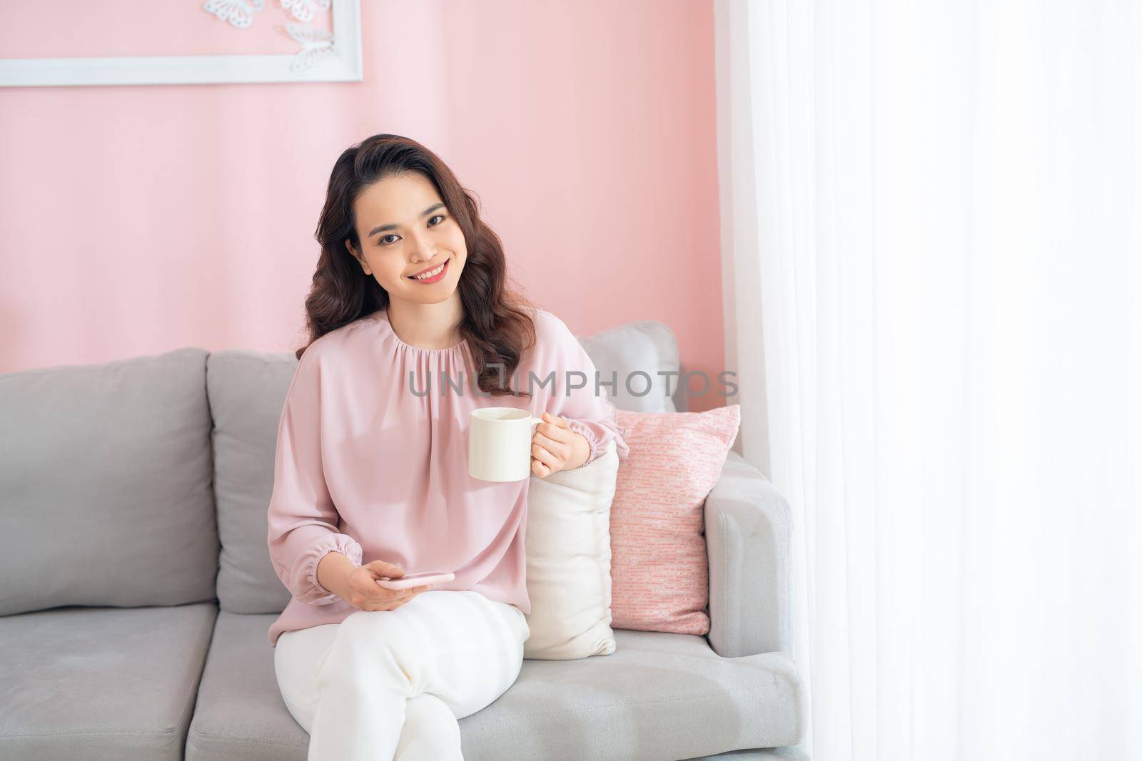 Attractive young Asian woman using phone and drinking coffee when sitting on sofa at home.