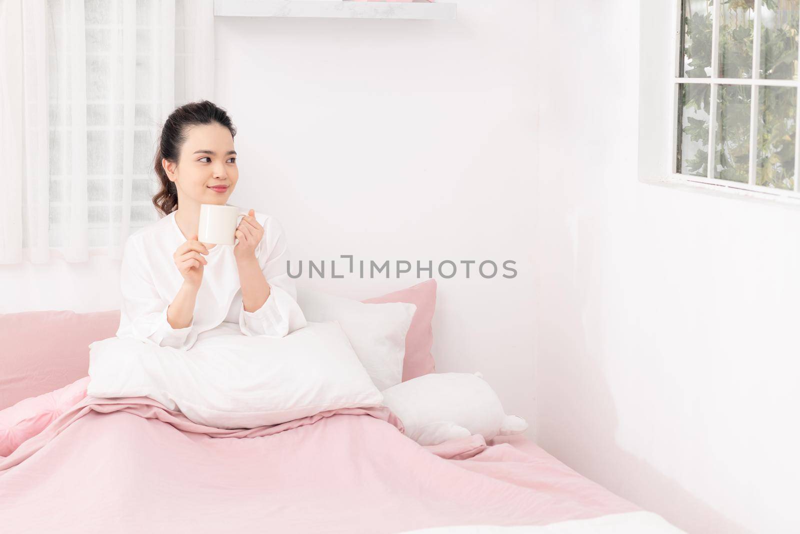 Relaxed young woman sitting on bed reading book and drinking coffee. Woman reading book on bed at home.