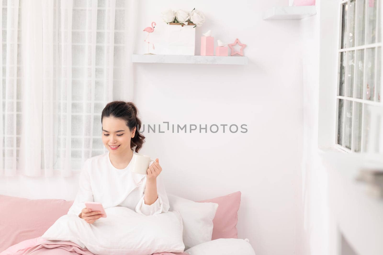 young woman student sitting on bed in her room drinking coffee and using mobile phone by makidotvn