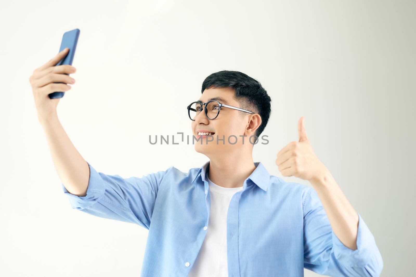 Cheerful young man is making selfie on the camera of his phone and showing thumb up