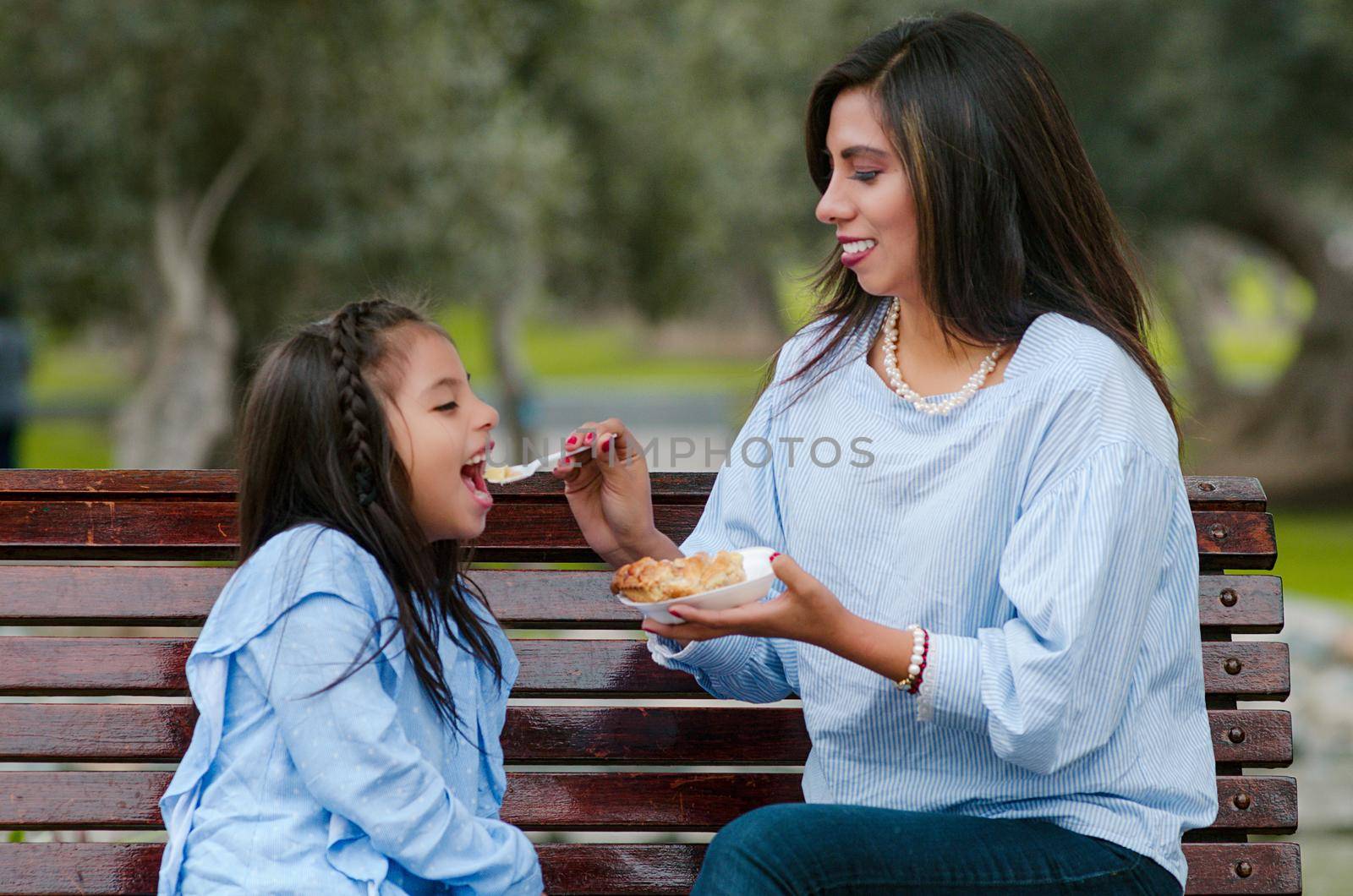 Mother and her little daughter sitting on a bench in the park eating a piece of cake