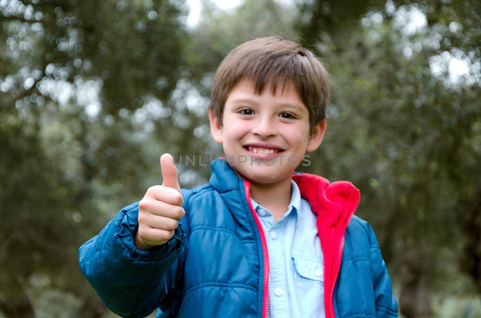 Portrait of a cute blond-haired boy, thumbs up and smile, with green background in the park