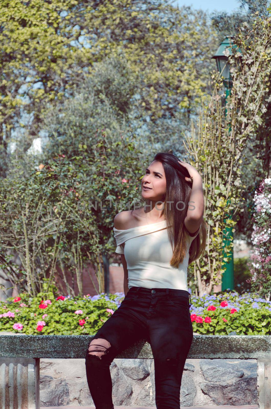 Happy young woman sitting on bench in city park Stylish fashion model wearing ripped jeans and pullover