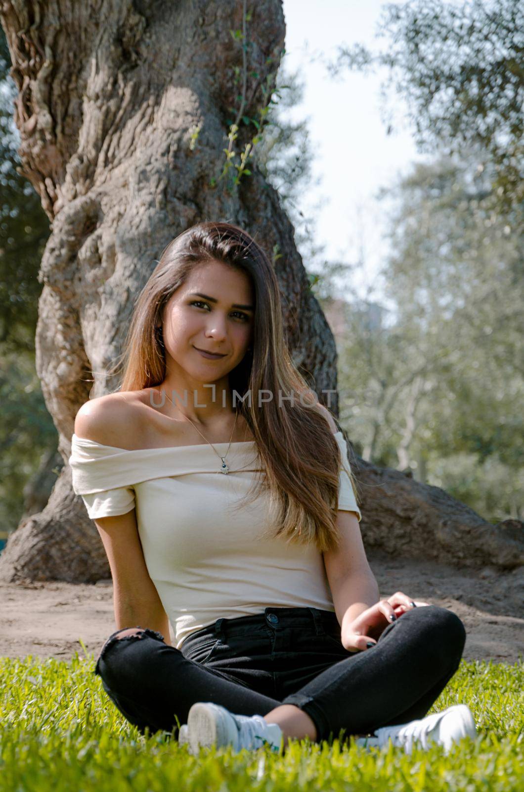 Stylish cheerful brunette sitting under a tree in a park on a sunny day