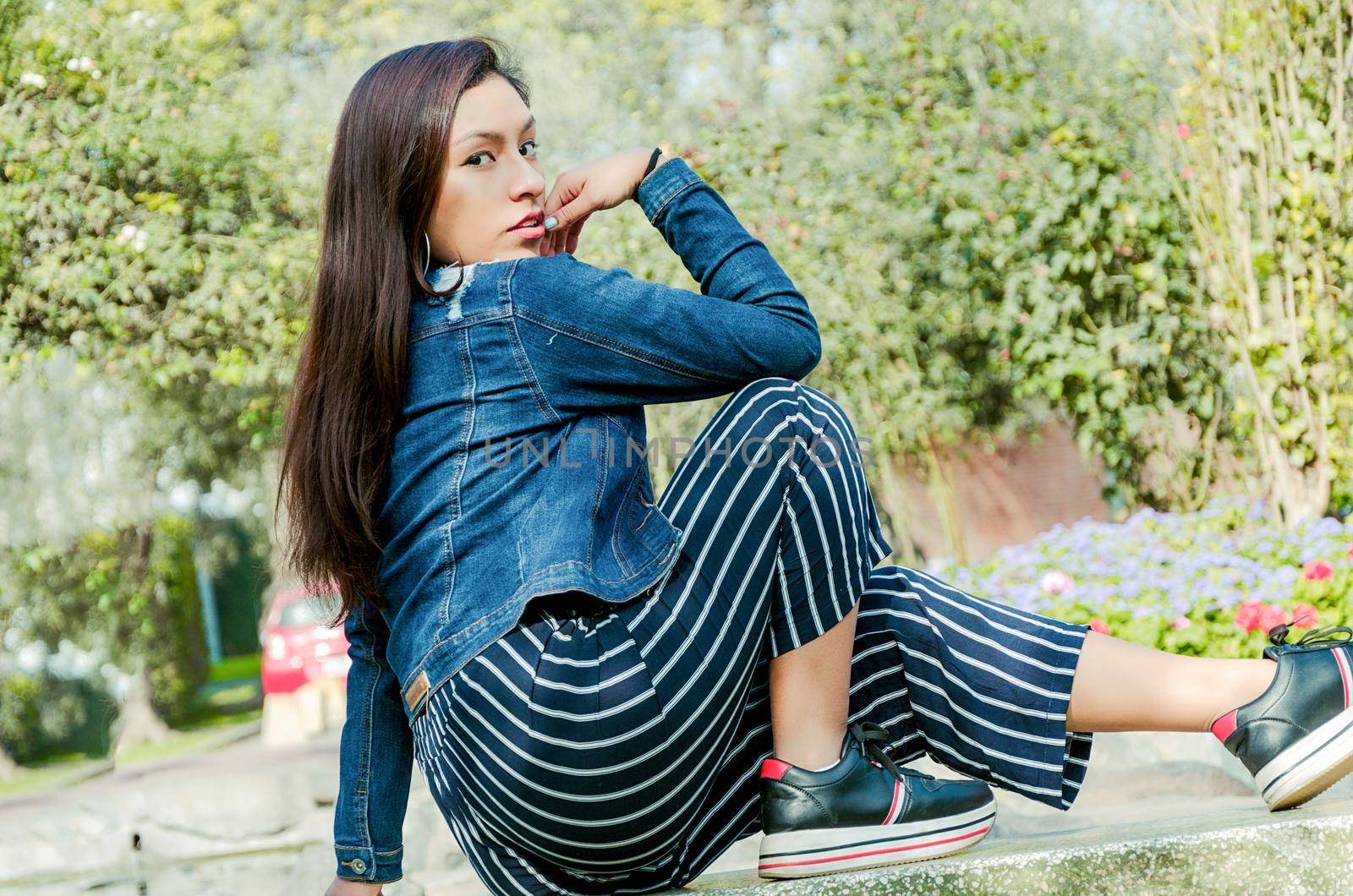 Outdoors lifestyle fashion portrait of pretty fashionable woman sitting on the marble bench. With an elegant wide jacket of jeans and black pants