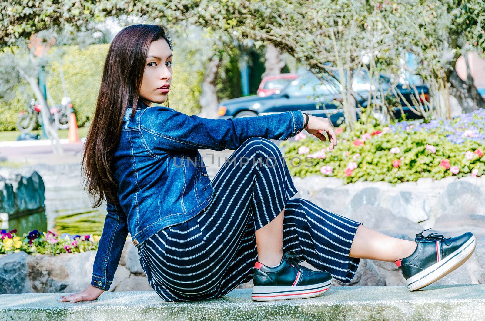 Outdoors lifestyle fashion portrait of pretty fashionable woman sitting on the marble bench. With an elegant wide jacket of jeans and black pants