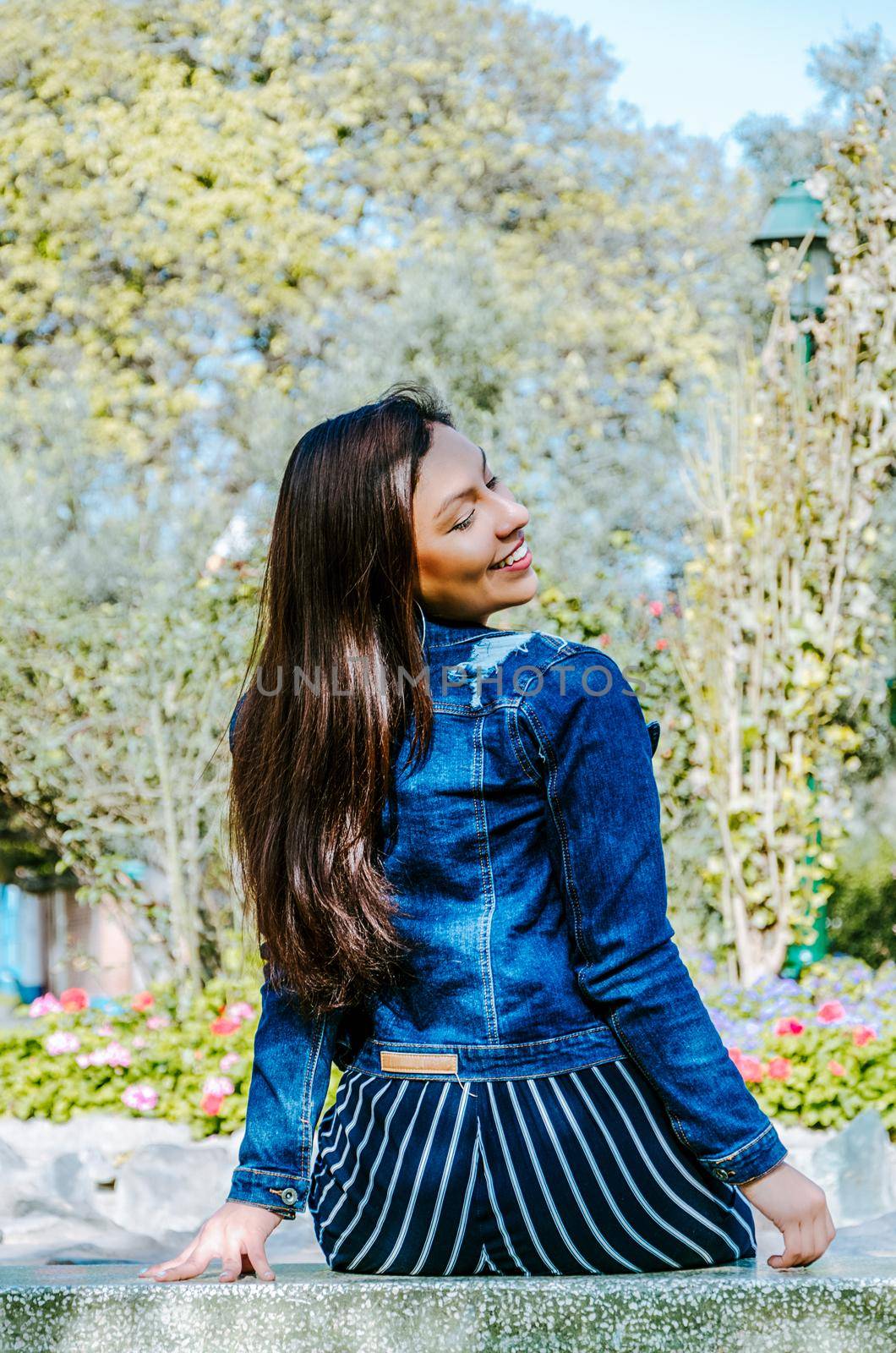 Outdoors lifestyle fashion portrait of pretty fashionable woman sitting on the marble bench. With an elegant wide jacket of jeans and black pants