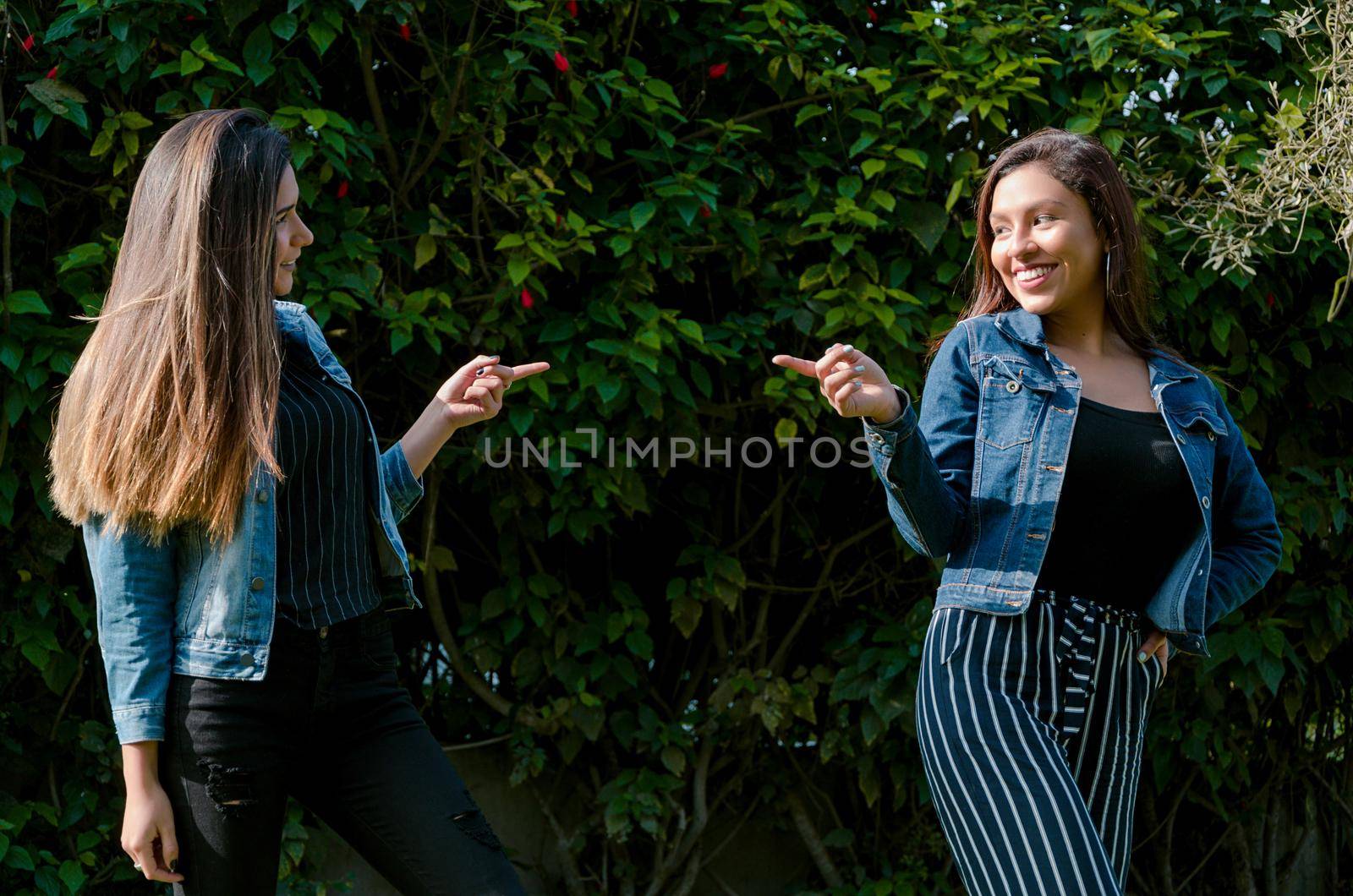 Cheerful friends are gossiping in the park. The girls are pointing with their finger and laughing. They wear fashionable clothes