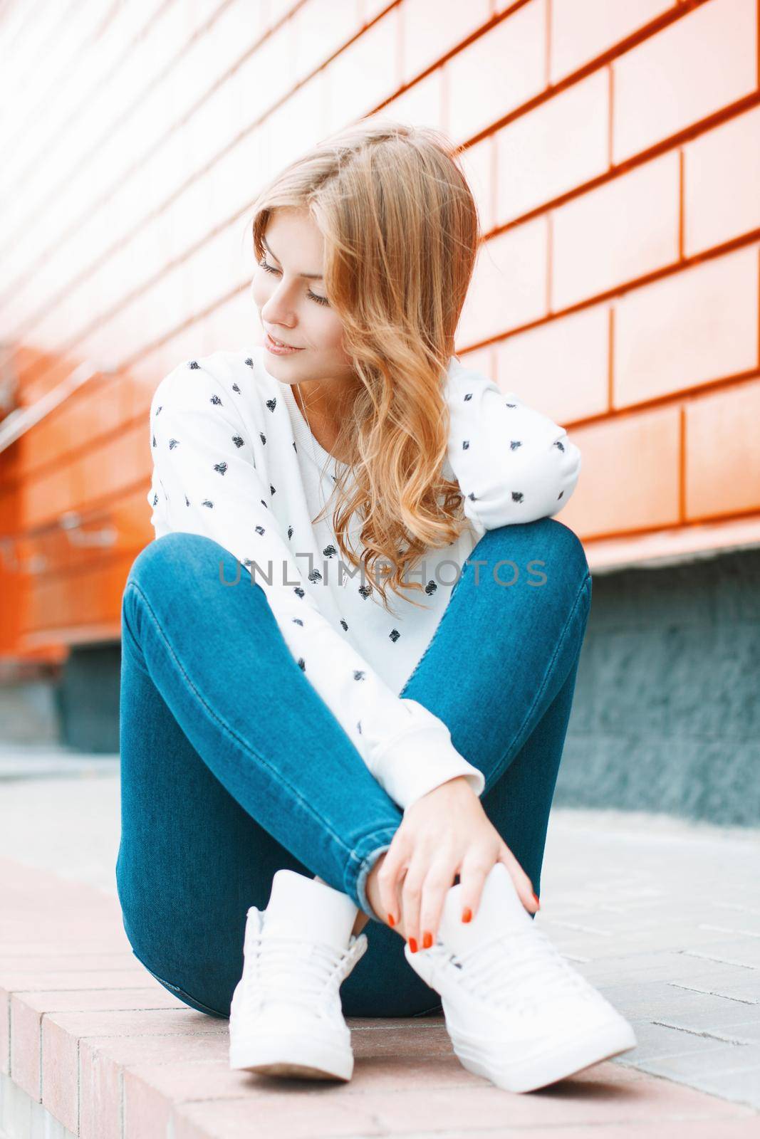 Beautiful woman in a white sweater, jeans and white sneakers sitting on a tile near the orange wall by alones