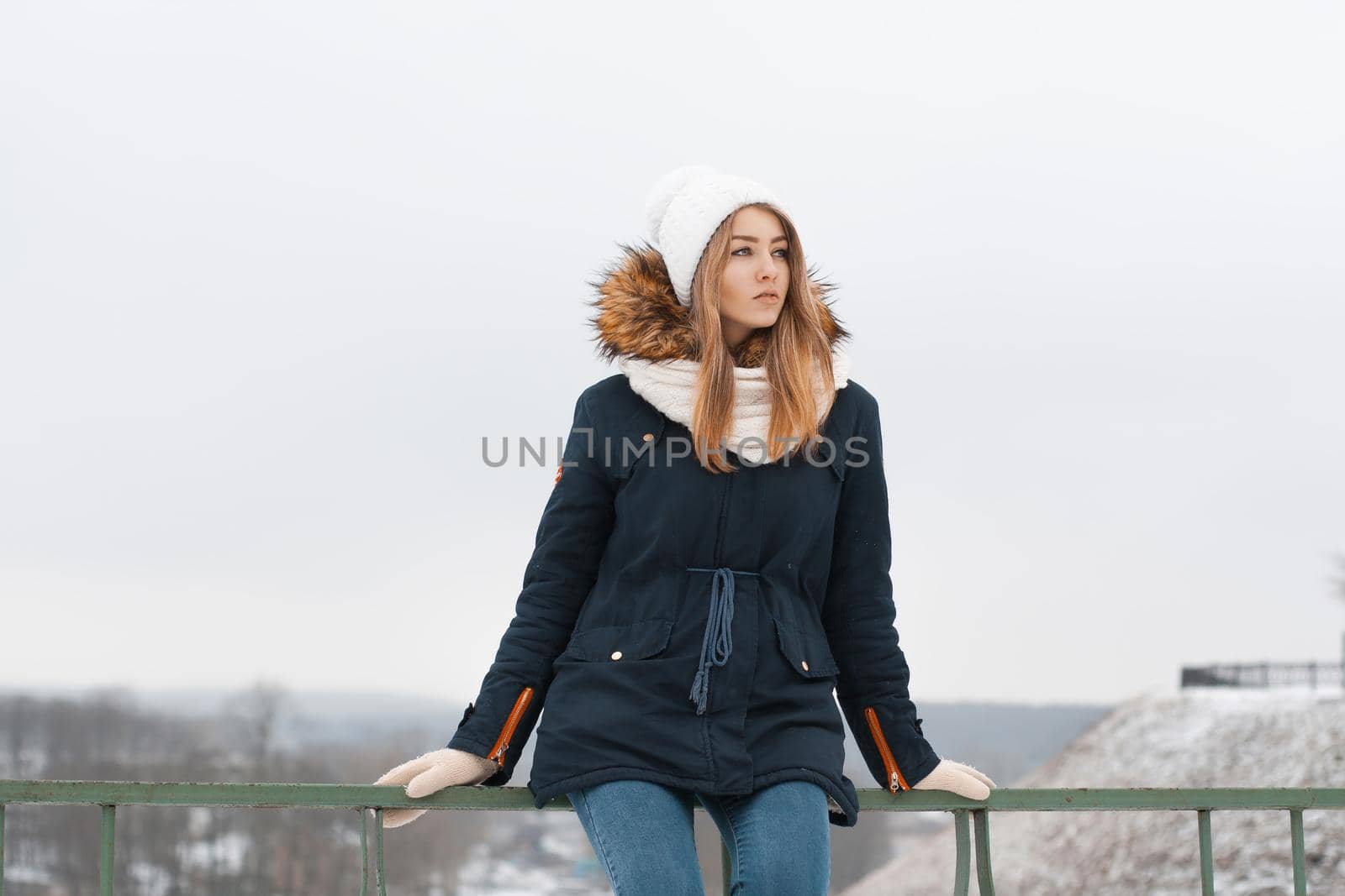 Cute girl in winter cap and jacket sitting on the railing. Outdoors in winter. by alones