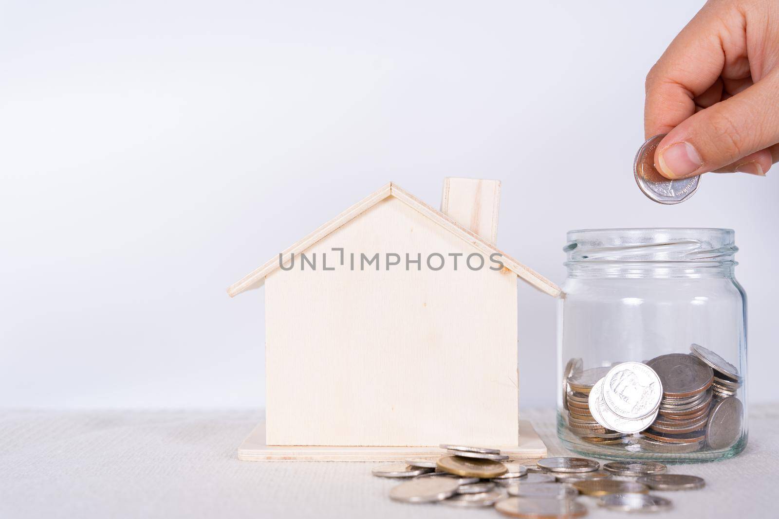 Hand put money coin into jar with wooden house isolated grey background. Saving money and investment concept.