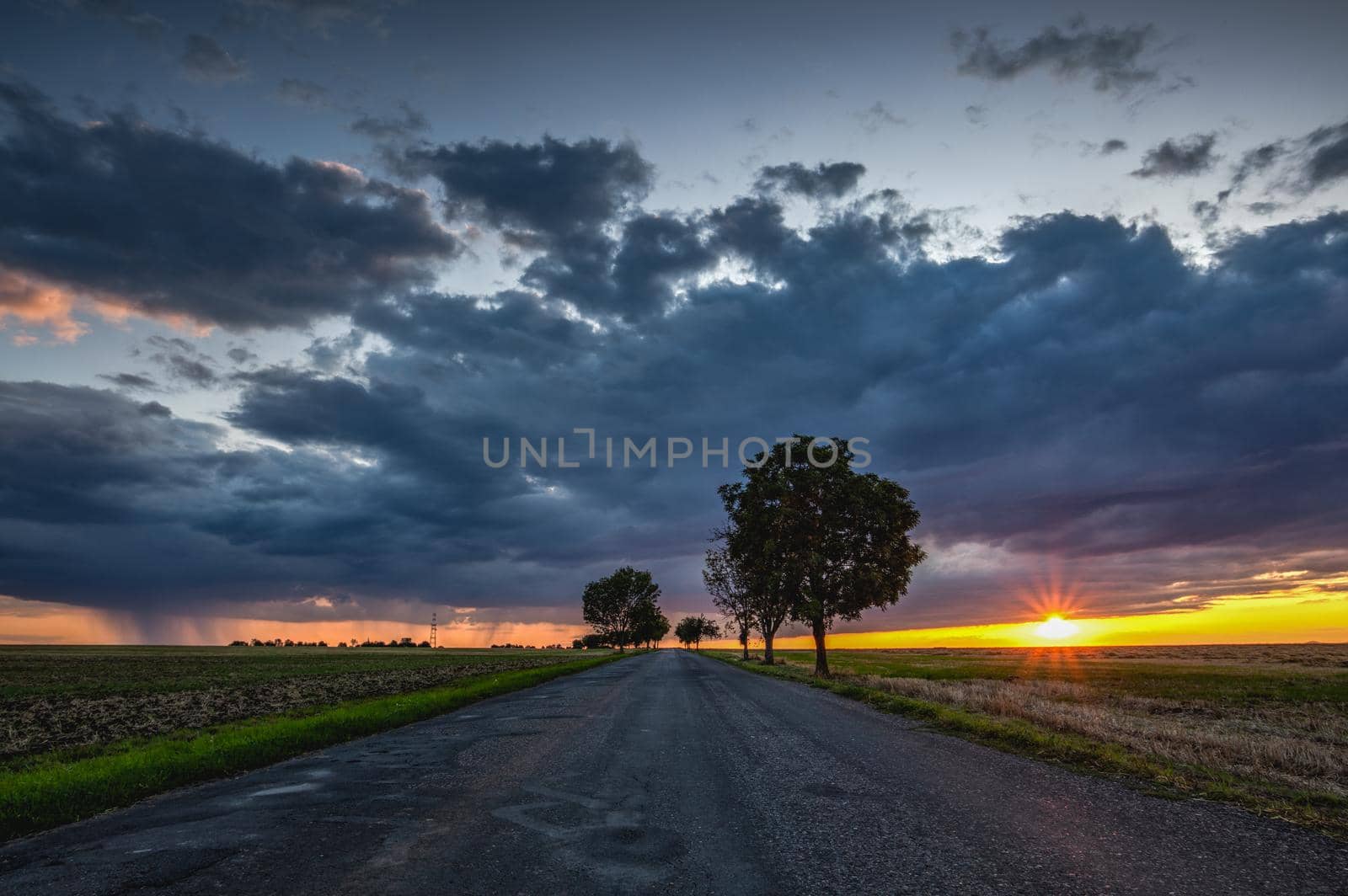 On the empty road between fields after harvesting at dramatic sunter. Picture with mowed wheat field  at sunset after rain. Czech Republic