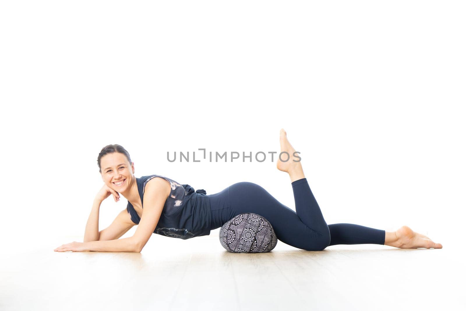 Restorative yoga with a bolster. Young sporty female yoga instructor in bright white yoga studio, lying on bolster cushion, stretching, smilling, showing love and passion for restorative yoga.
