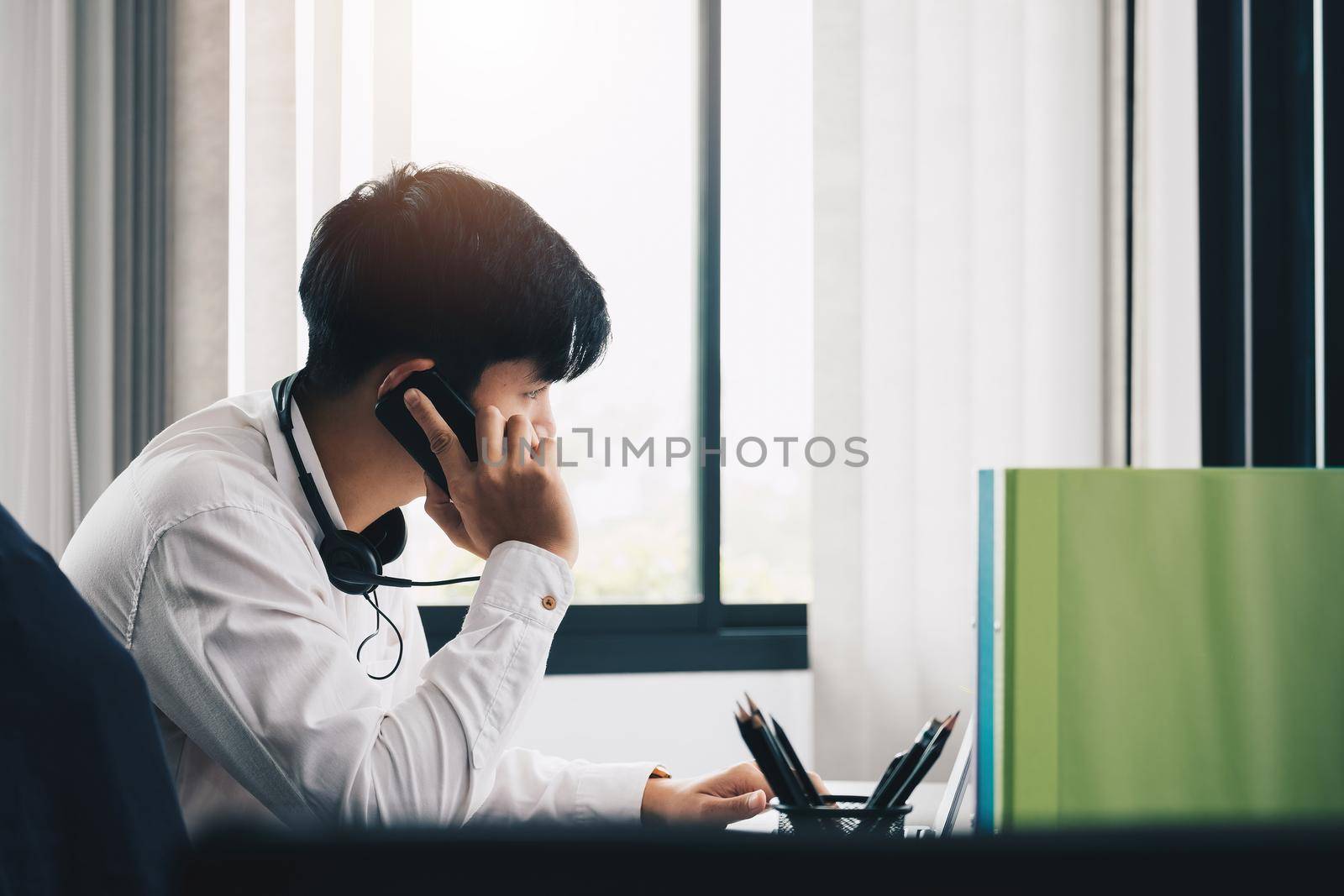 Side view of young man entrepreneur in office making phone call while working with laptop. by nateemee