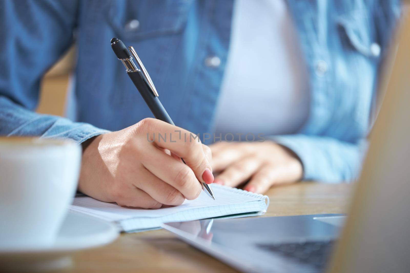 Close up of woman holding pen and writing some interesting information in notebook. Concept of process preparing for communication online with tasty coffee and modern laptop. 