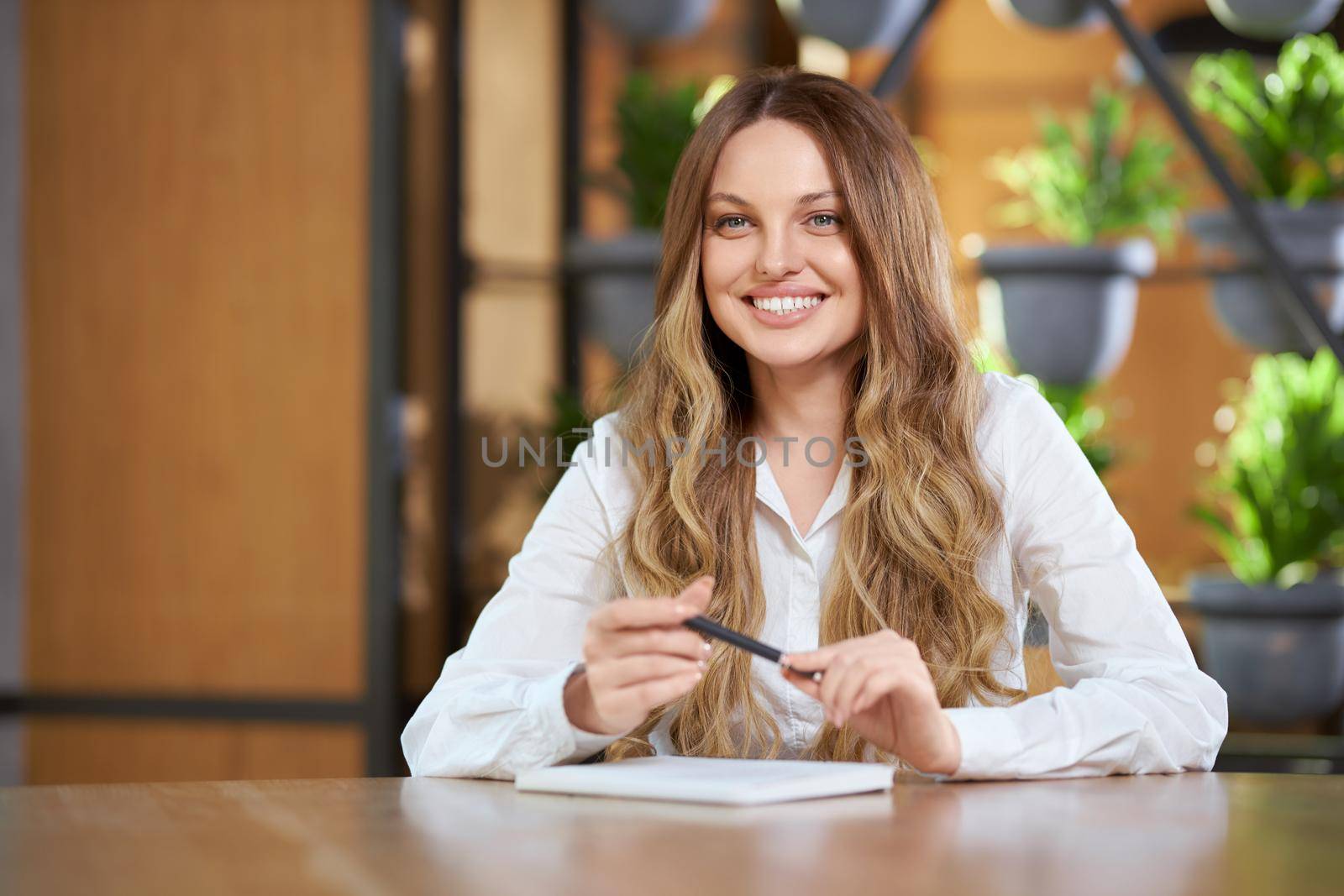 Close up portrait of smiling young woman with long hair in white shirt sitting at the table and writing different information. Concept of process interview or communicating in cafe. 