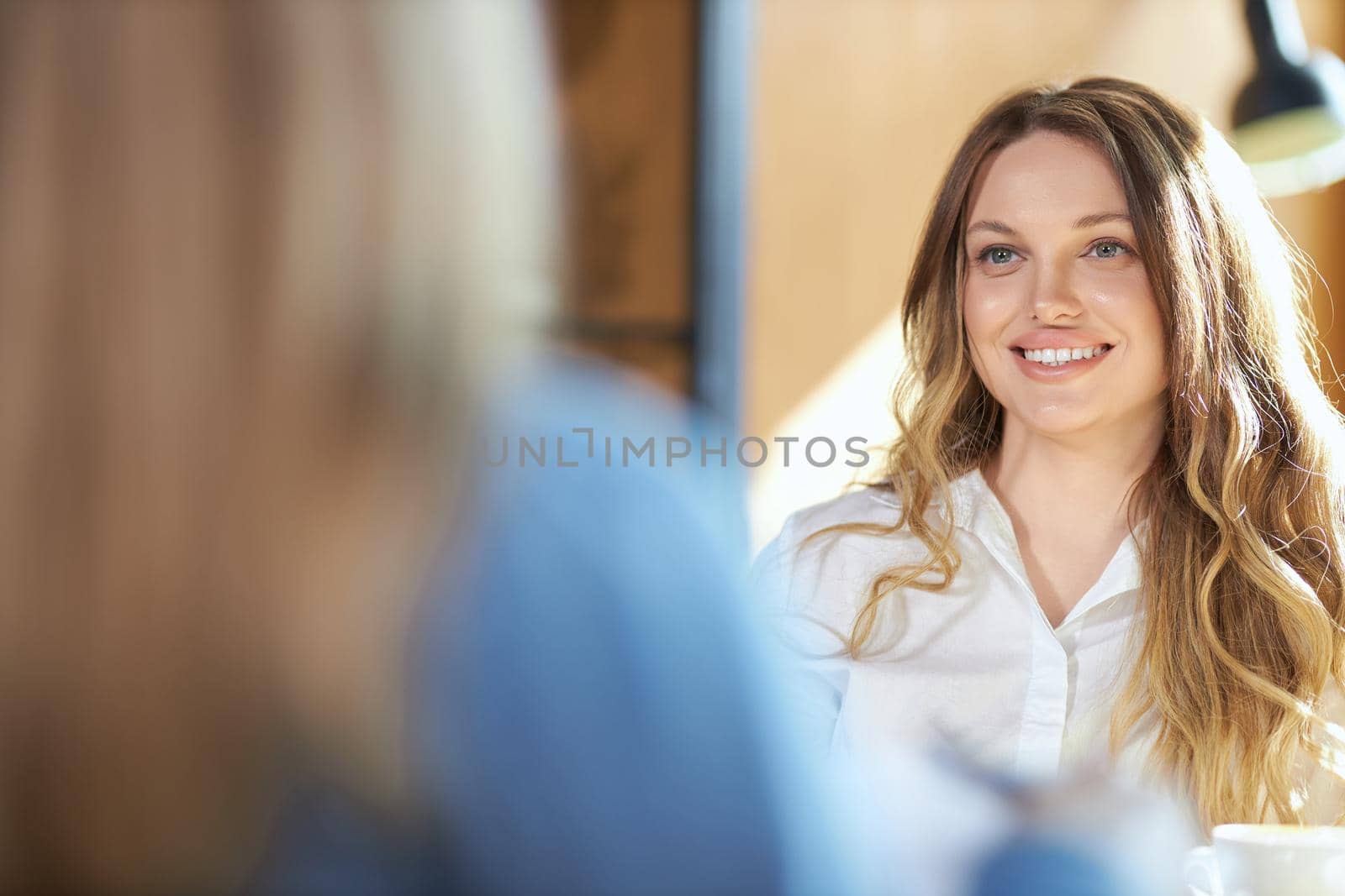 Close up of smiling cute young woman in white shirt sitting at the table with delicious coffee and communicating with friend. Concept of process talking about different topics. 