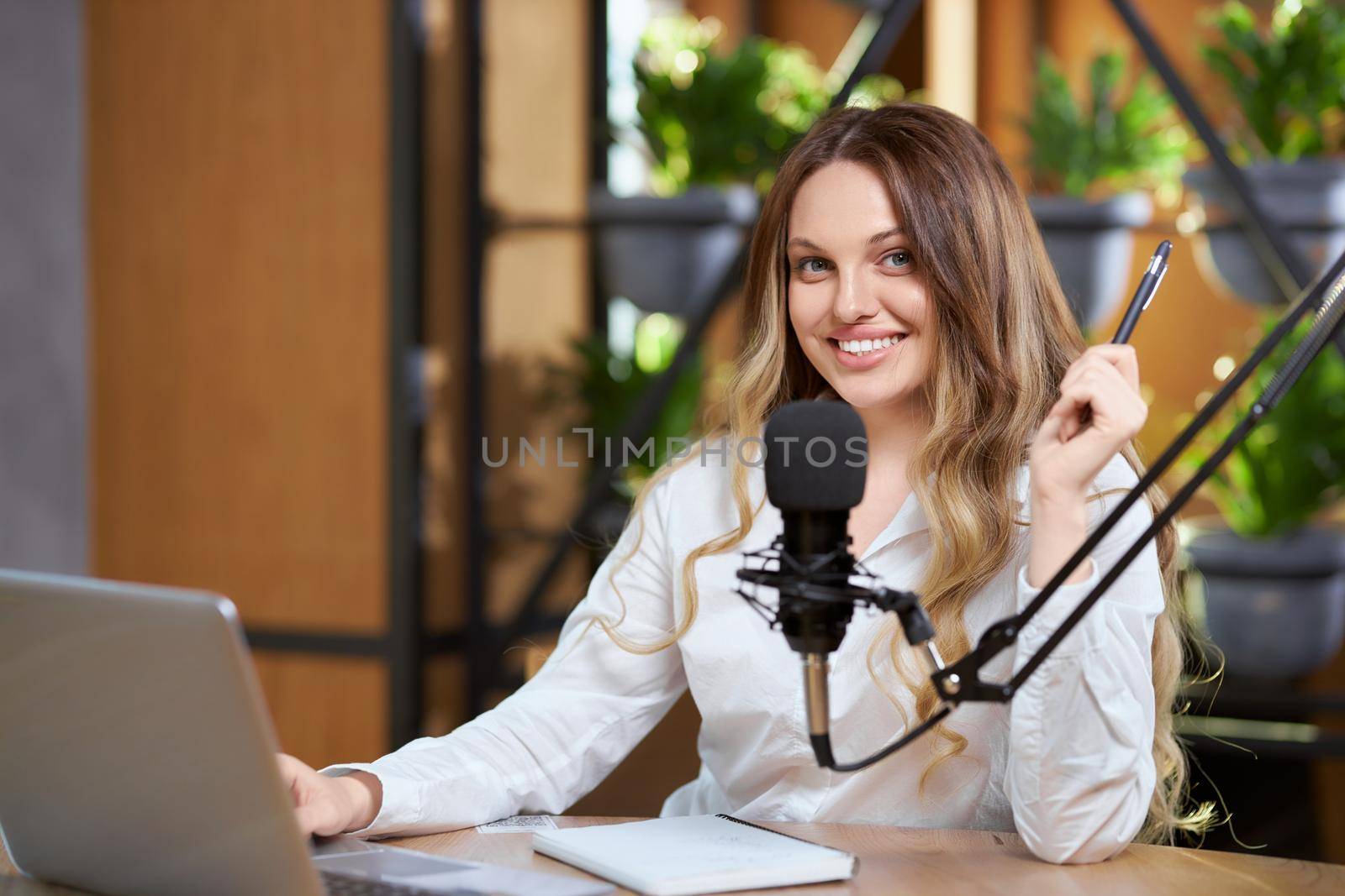 Close up of smiling blogger woman in white shirt sitting at the table and communicating with followers online by laptop. Concept of process live broadcast with special modern microphone. 