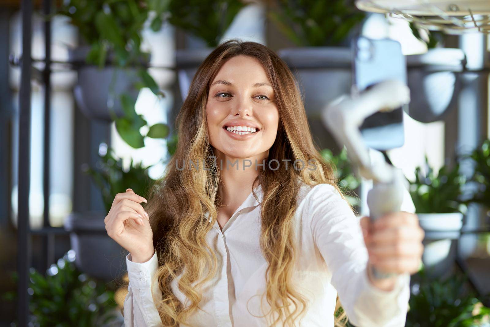 Front view portrait of smiling attractive young woman in white shirt holding selfie stick and communicating with people in live broadcast. Concept of process doing video or selfie in cafe. 