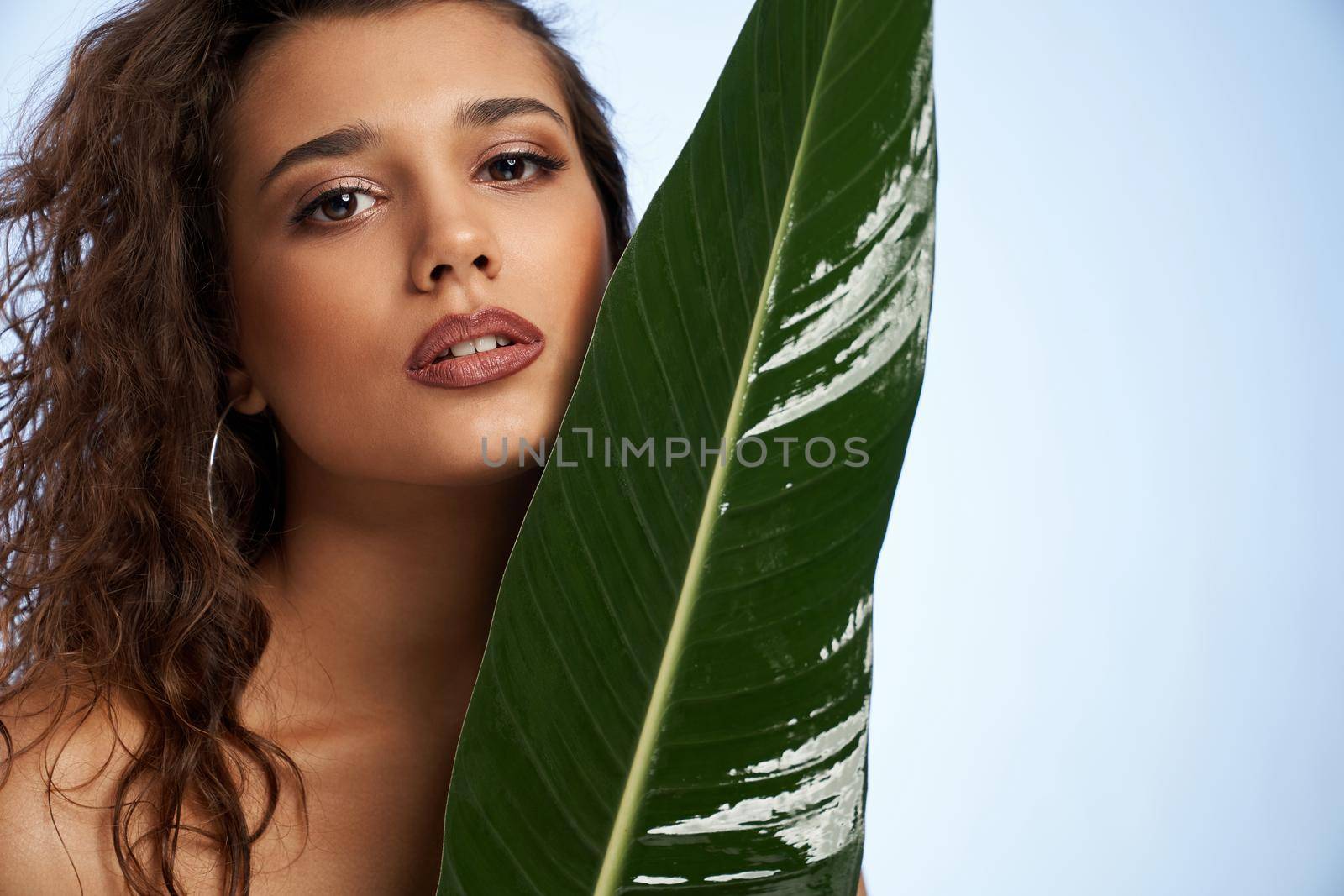 Portrait of beautiful model with curly hair with big green leaf on light blue background. Concept of shooting with green plants for fashion magazine on professional studio.