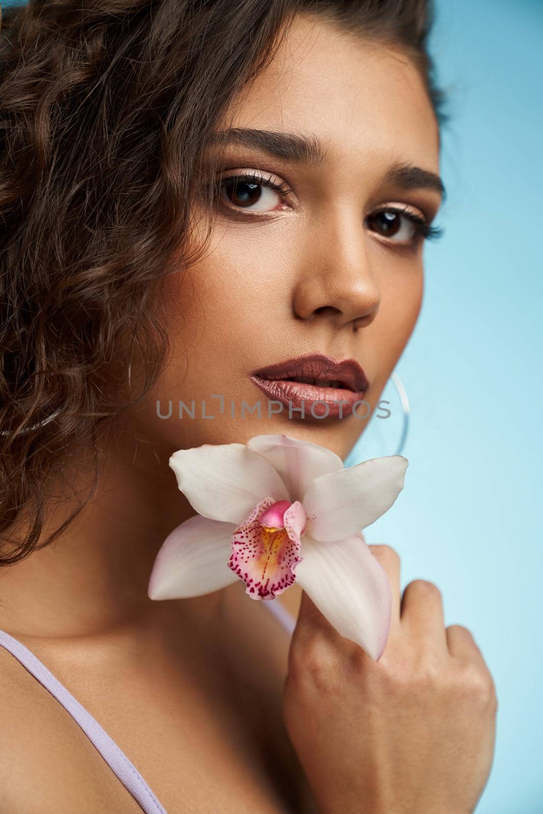Portrait of attractive model with trendy makeup and curly hair holding beautiful white orchid on blue background. Concept of sensual and tender photo shoot on studio.